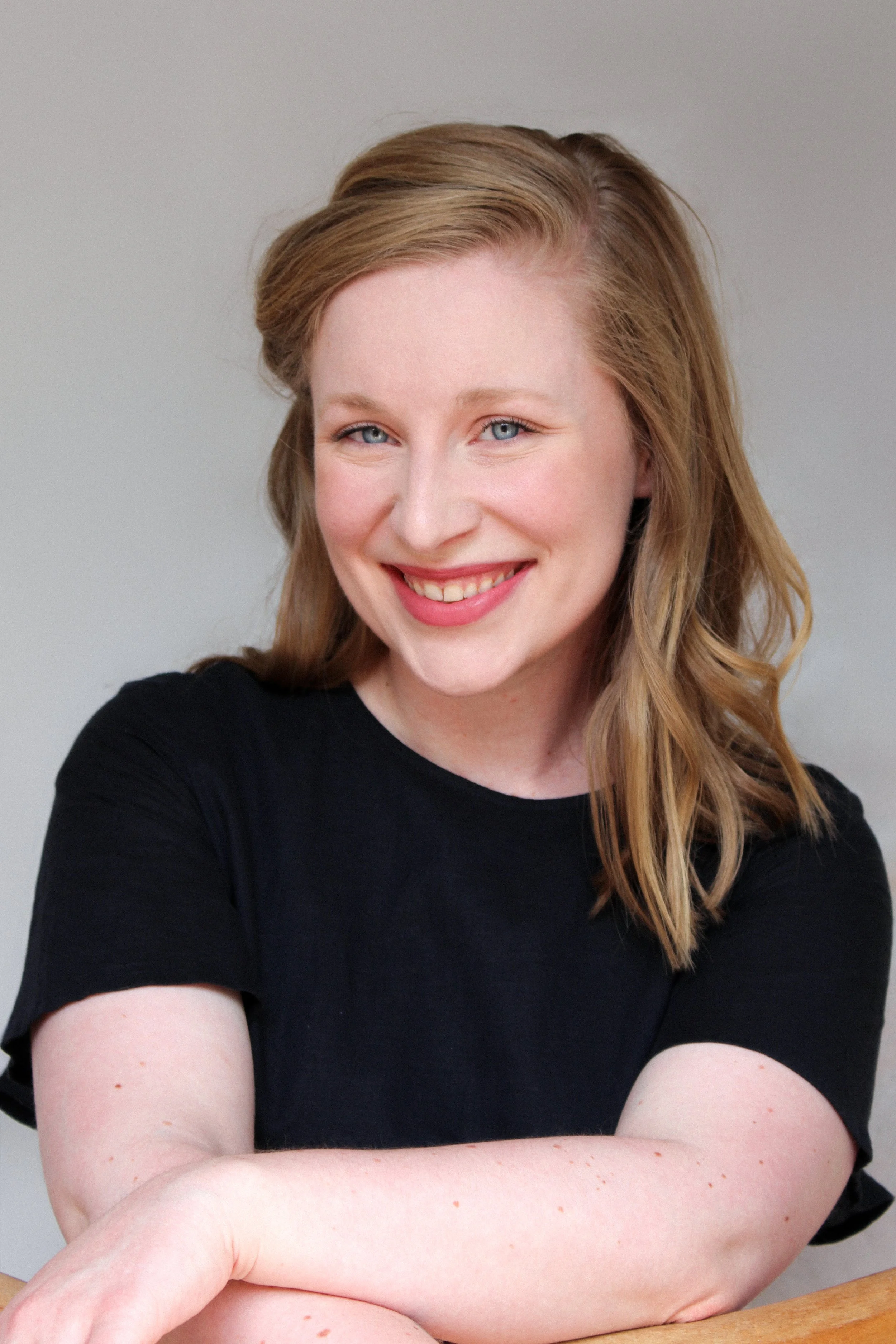 A woman with red hair and blue eyes, smiling at the camera, wearing a black shirt, with her arms crossed.
