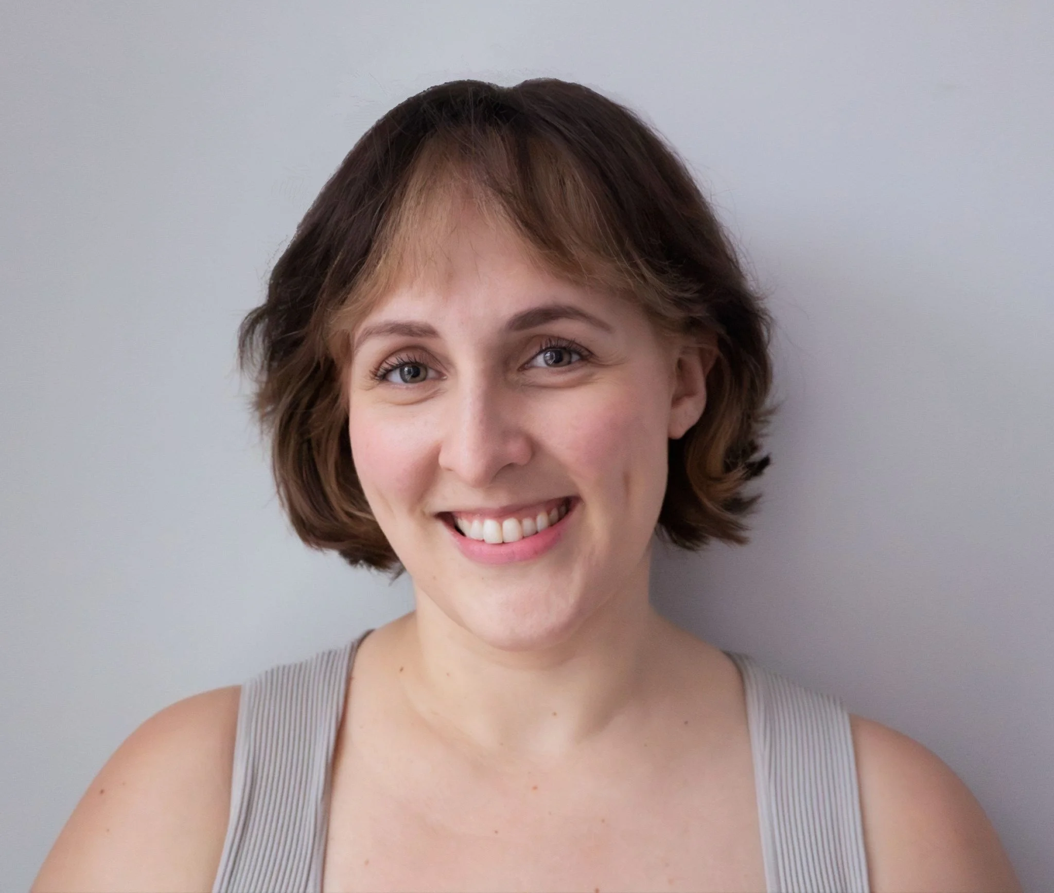 Close-up portrait of a smiling woman with short brown hair, wearing a light gray sleeveless top, against a plain light background.