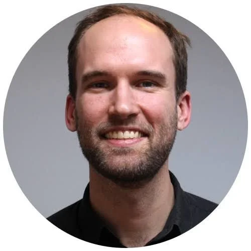 A smiling man with short brown hair, face beard, wearing a black shirt against a gray background.