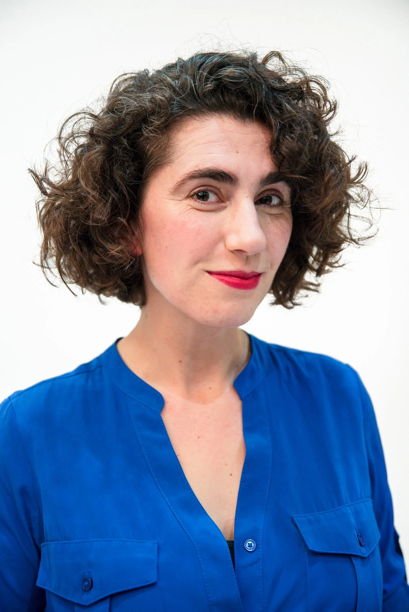 A woman with curly short brown hair, wearing a blue shirt, smiling slightly with red lipstick, against a white background.