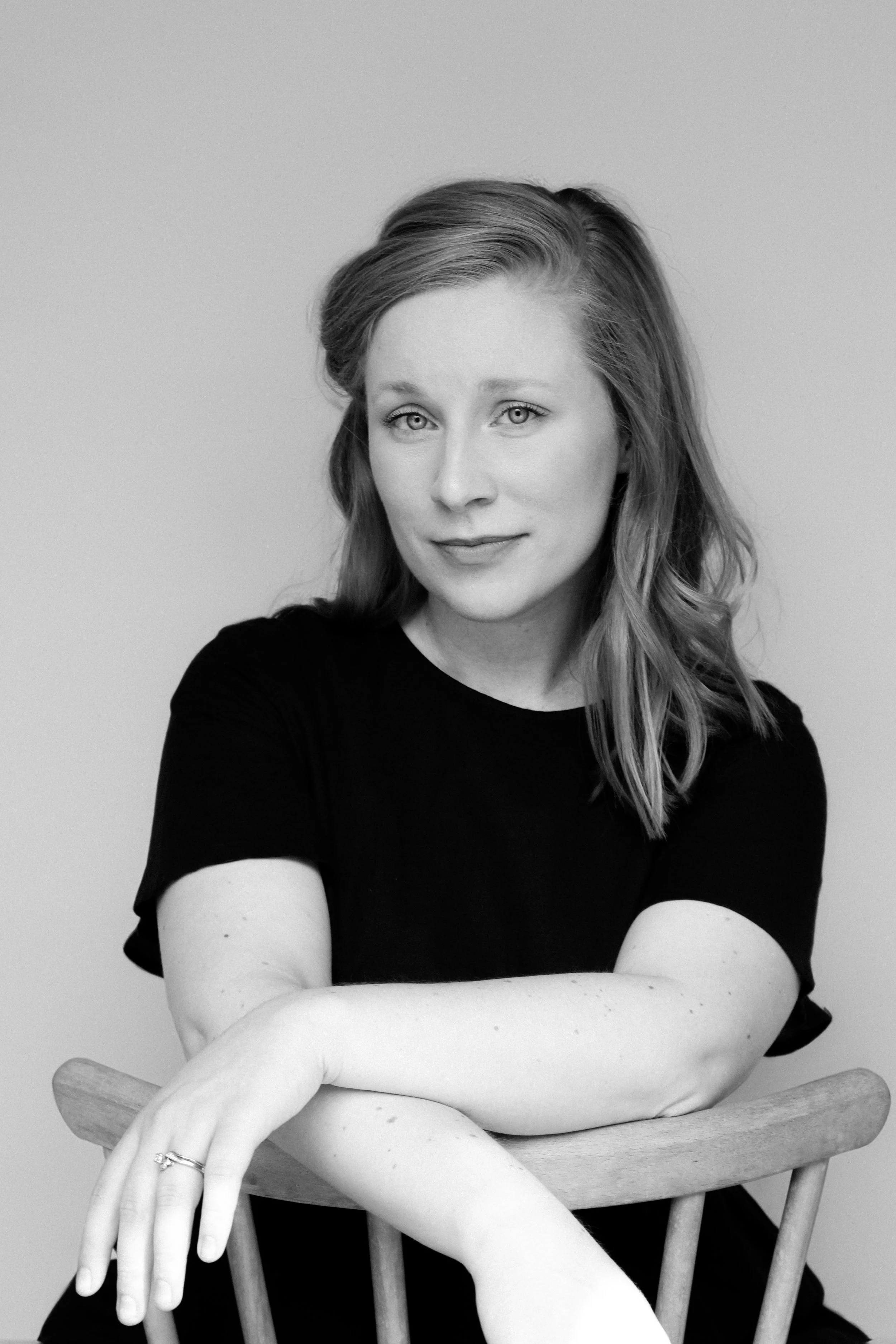 A black-and-white portrait of a woman with light hair, looking at the camera with a slight smile, sitting with her arms resting on the back of a wooden chair against a plain background.
