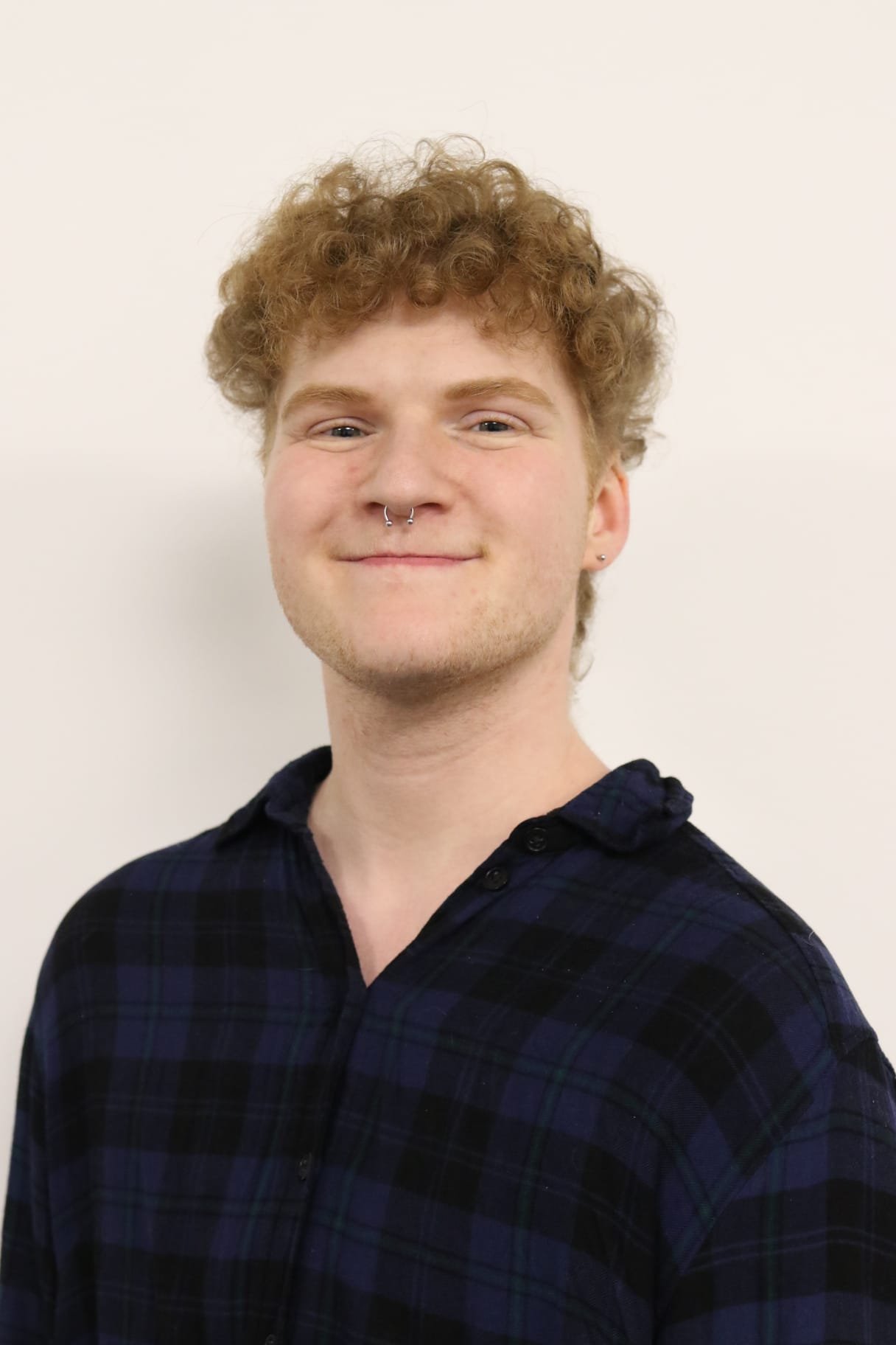 A young man with curly red hair, light skin, and a septum piercing, wearing a dark plaid shirt, standing against a plain white wall, smiling slightly.
