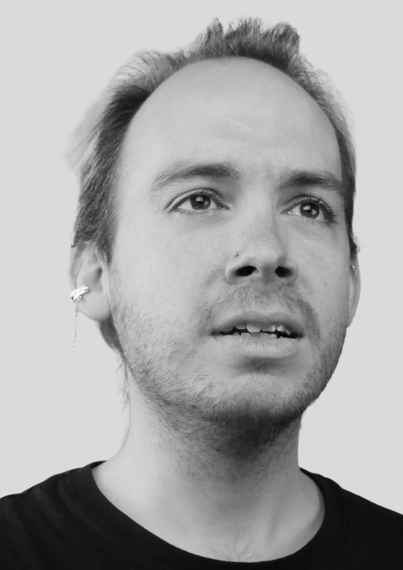 Close-up black and white photo of a young person with short hair, facial piercings, and earrings, looking slightly upward.