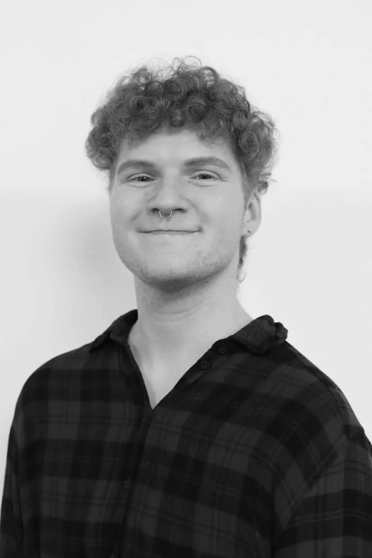 Black and white photo of a young person with curly hair, a septum piercing, and a small earring, smiling slightly, wearing a dark plaid shirt, standing against a plain light background.