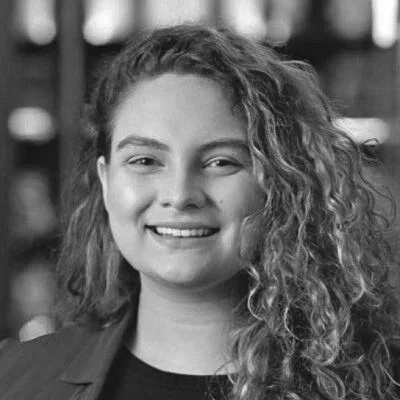 A young woman with curly hair smiling at the camera, in front of a blurry background.