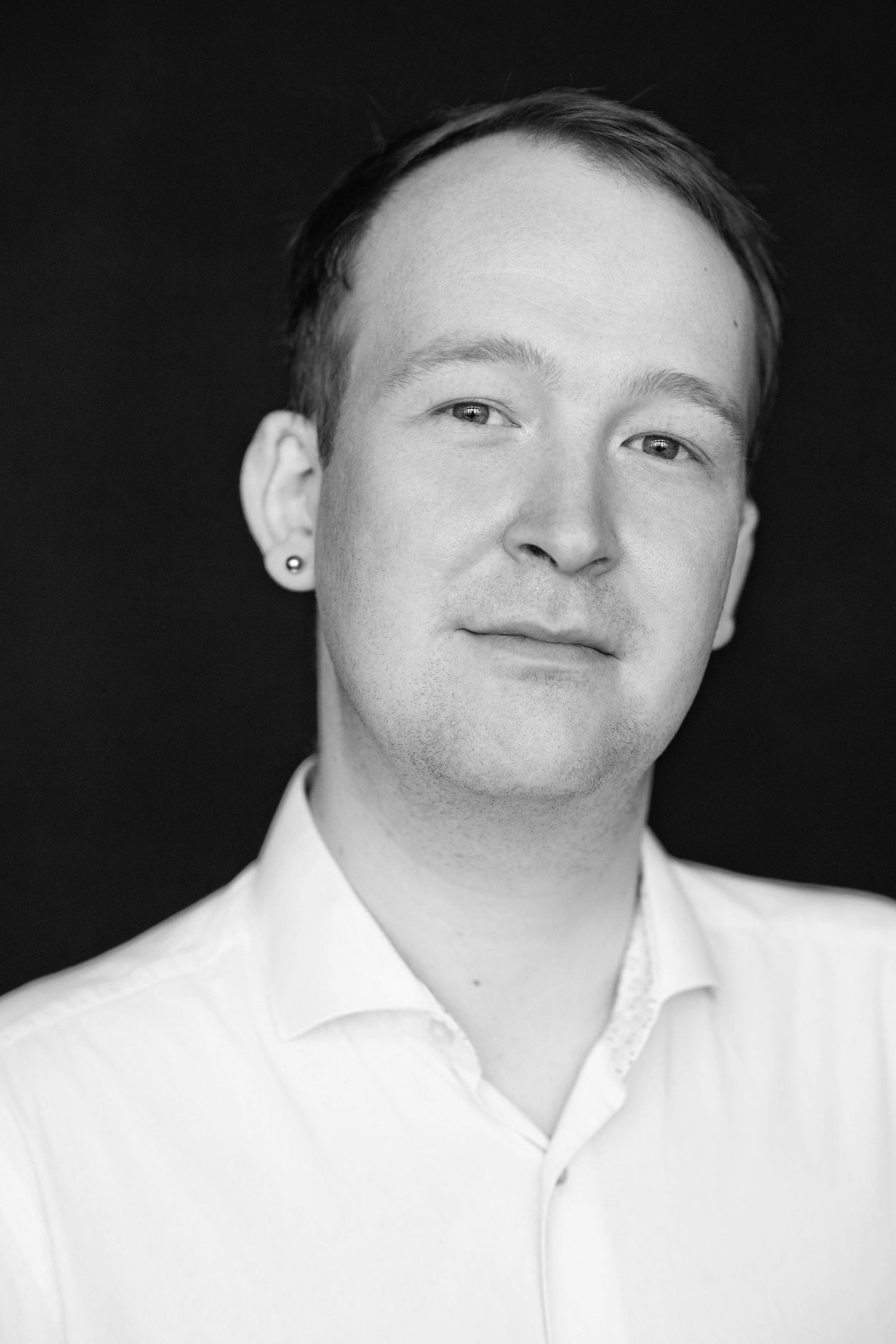 A young man smiling, wearing a suit and tie, sitting indoors against a modern architectural background in black and white.