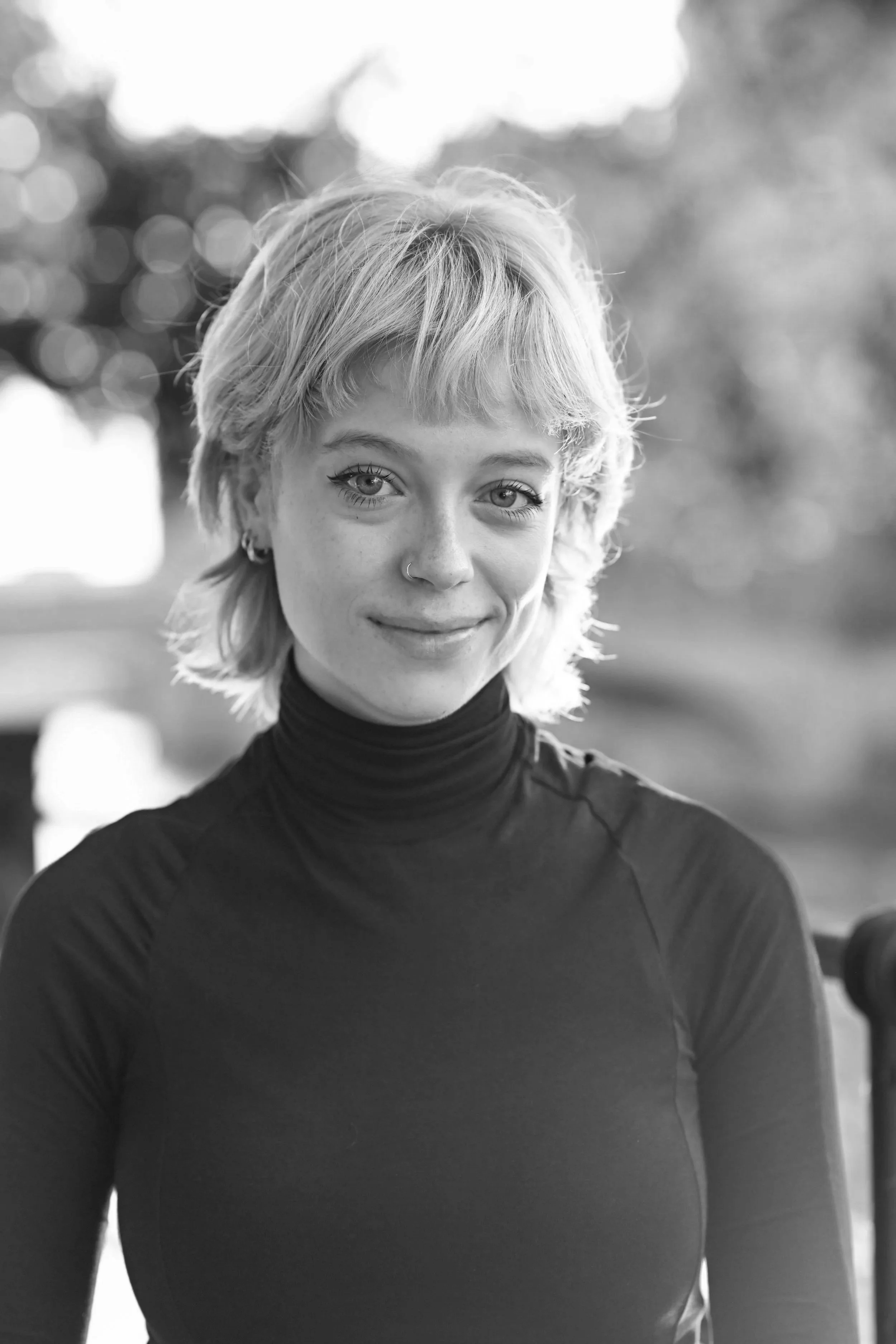 A black and white portrait of a young woman with short, wavy, blonde hair, wearing a high-neck black top, smiling softly at the camera outdoors.