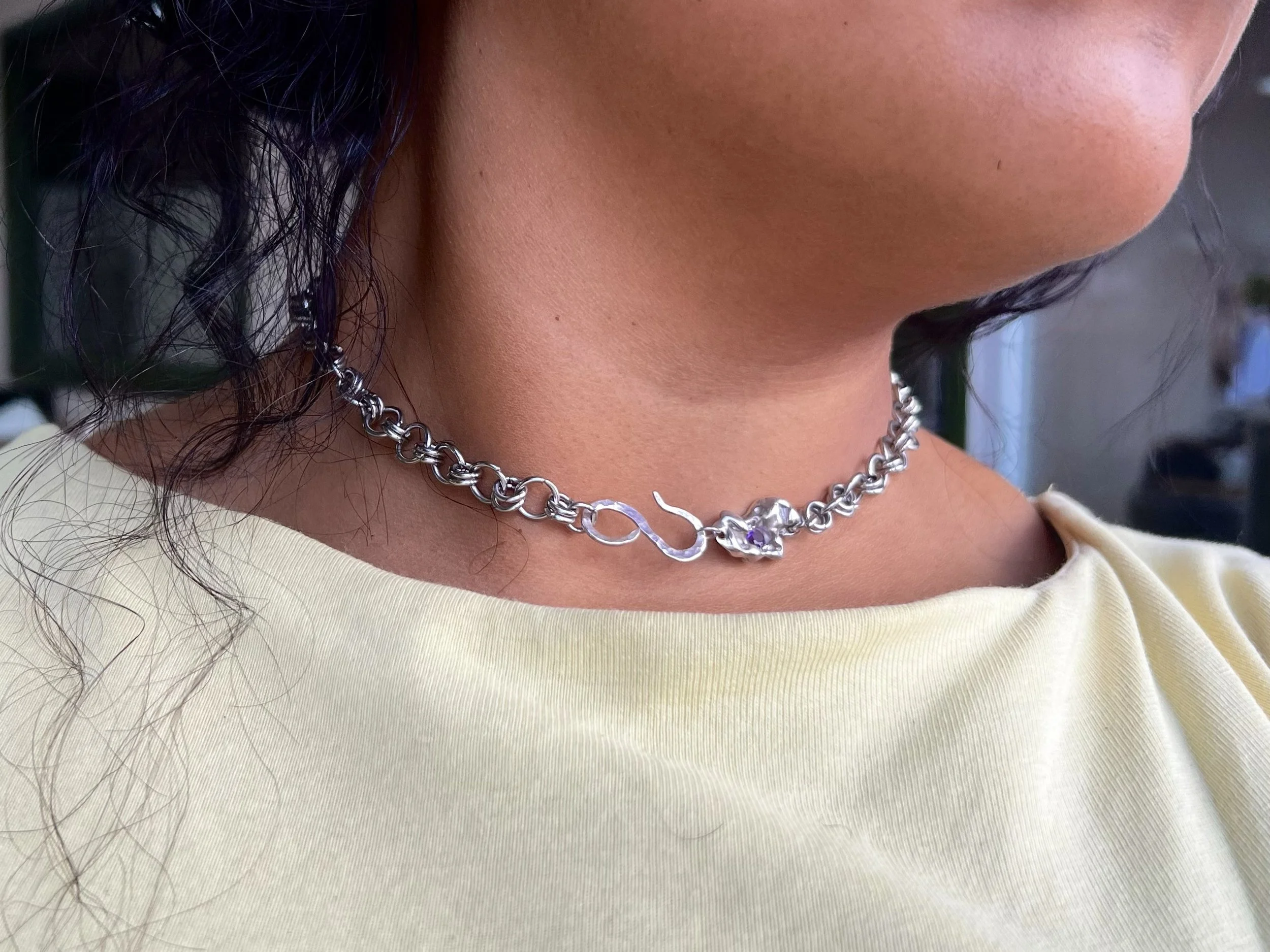 Close-up of a person's neck and a portion of their face, showing dark, curly hair and wearing a silver chain necklace, with a light-colored shirt or top.