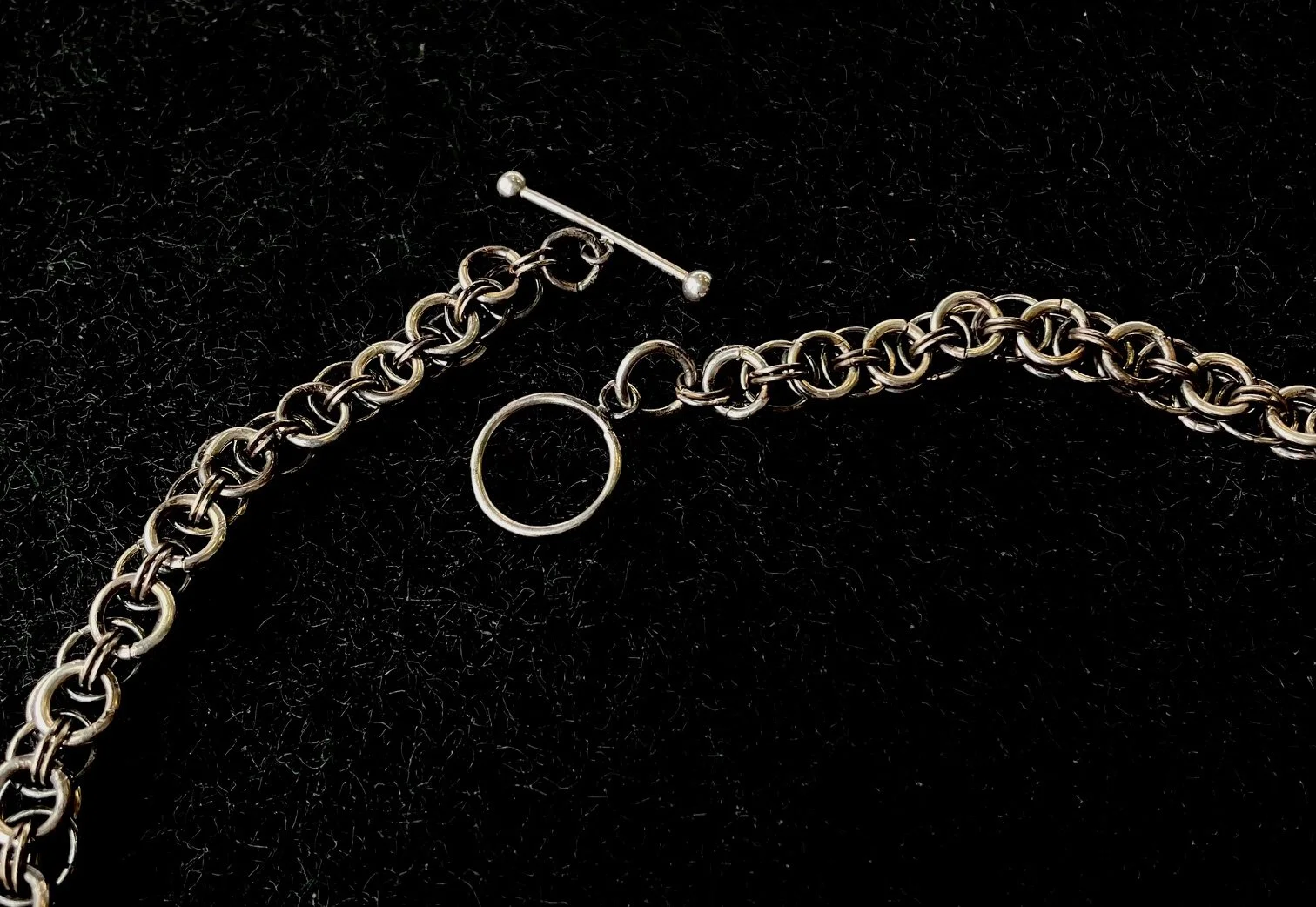 Silver jewelry chain, ring, and bar clasp on black fabric background.