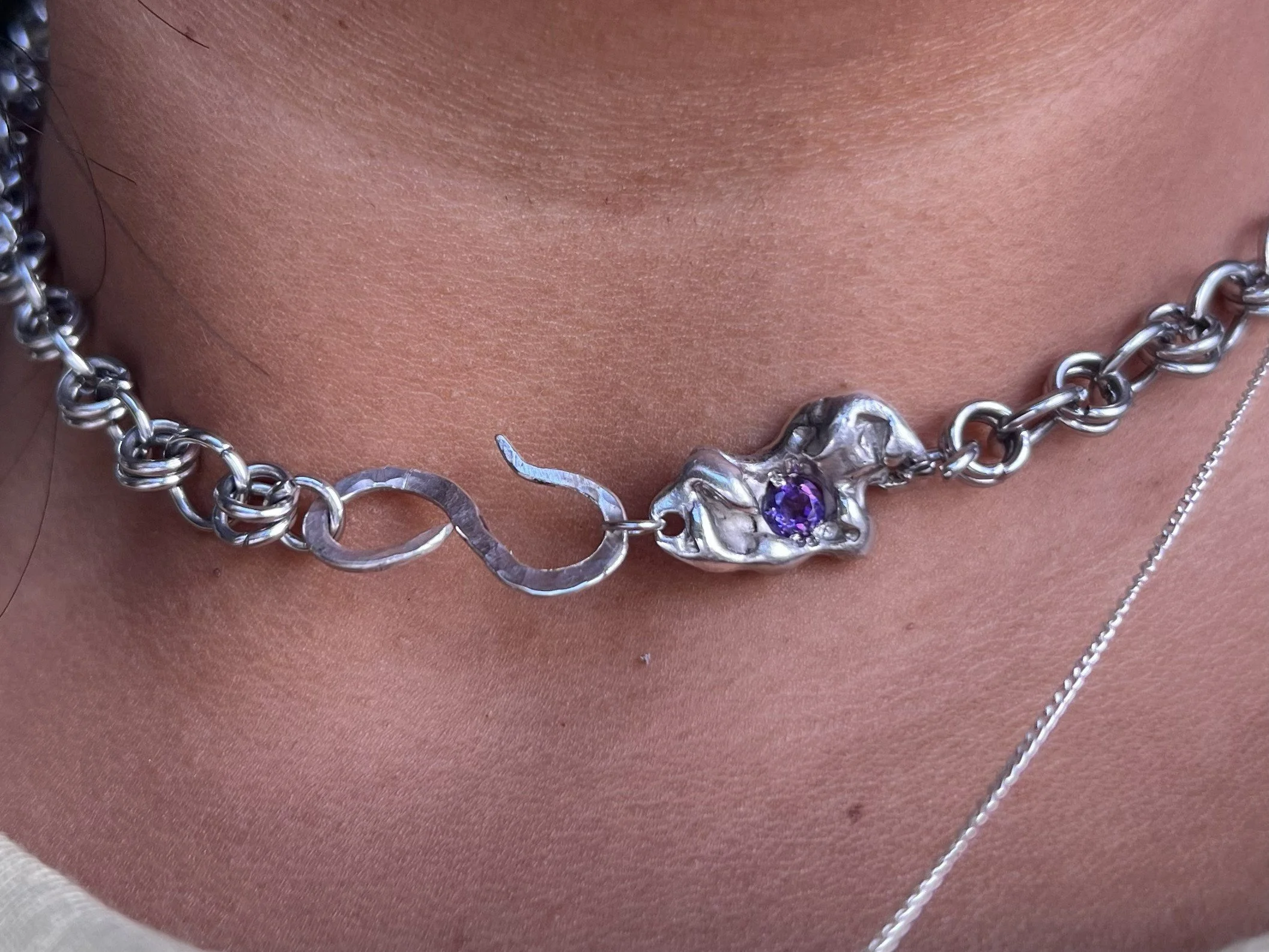 Close-up of a person's neck and chest showing a silver chain necklace with purple gemstones and a silver pendant, with a hint of a white shirt strap on the bottom left.