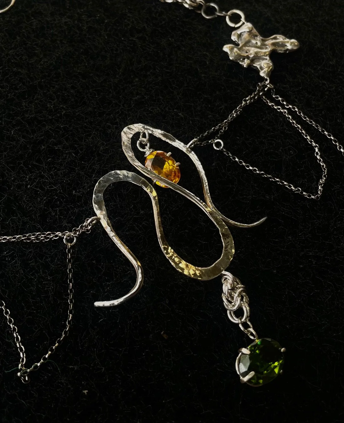 Silver necklace with a pendant shaped like a swan, featuring an amber-colored stone and a green gemstone hanging from its tail, on a black background.