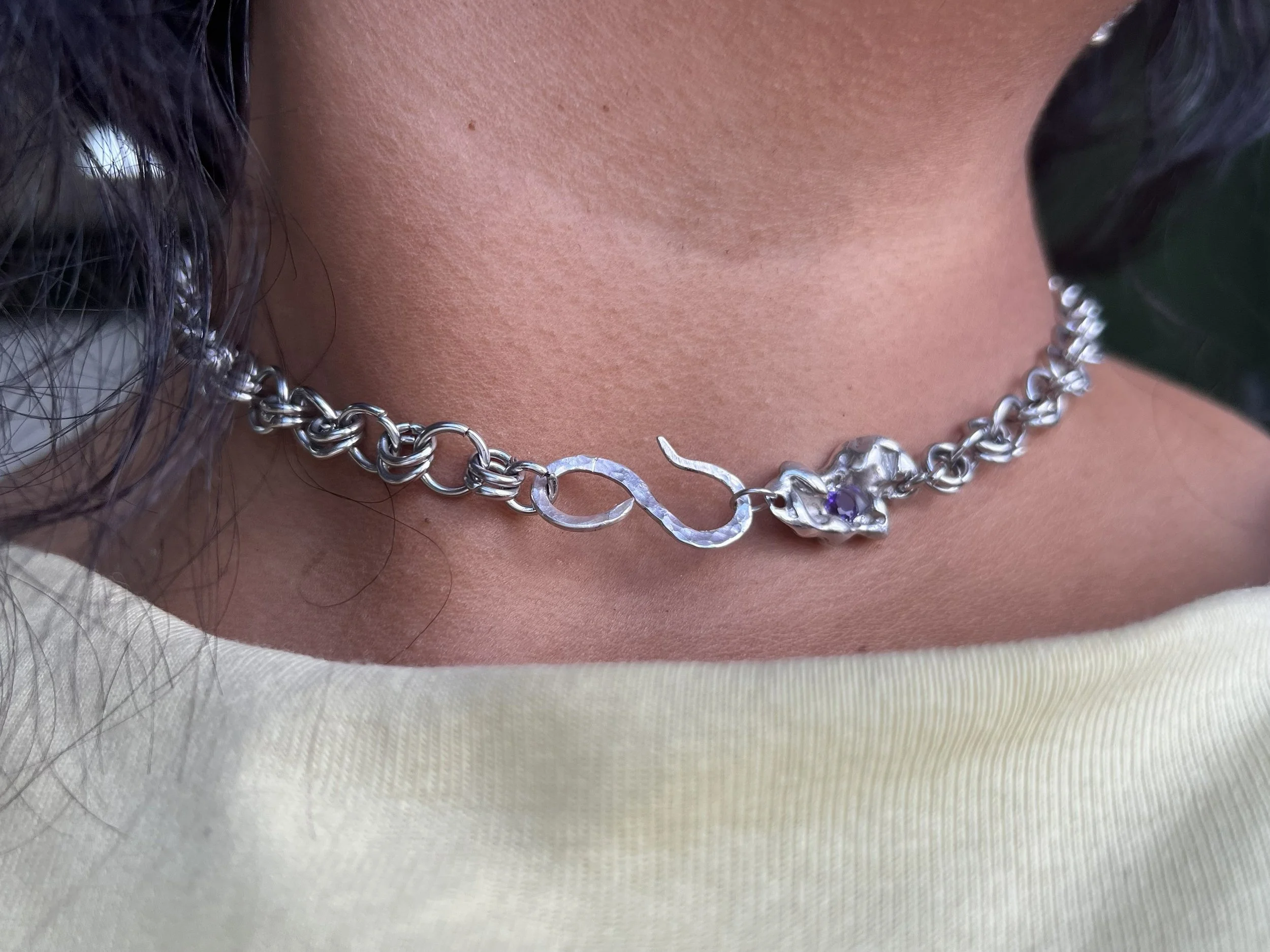 Close-up of a person's neck and collarbone area showing a silver chain necklace with a signature-shaped pendant and a small purple gemstone. Part of dark hair and a dark earring are visible on the left side, and the person is wearing a light-colored fabric top.