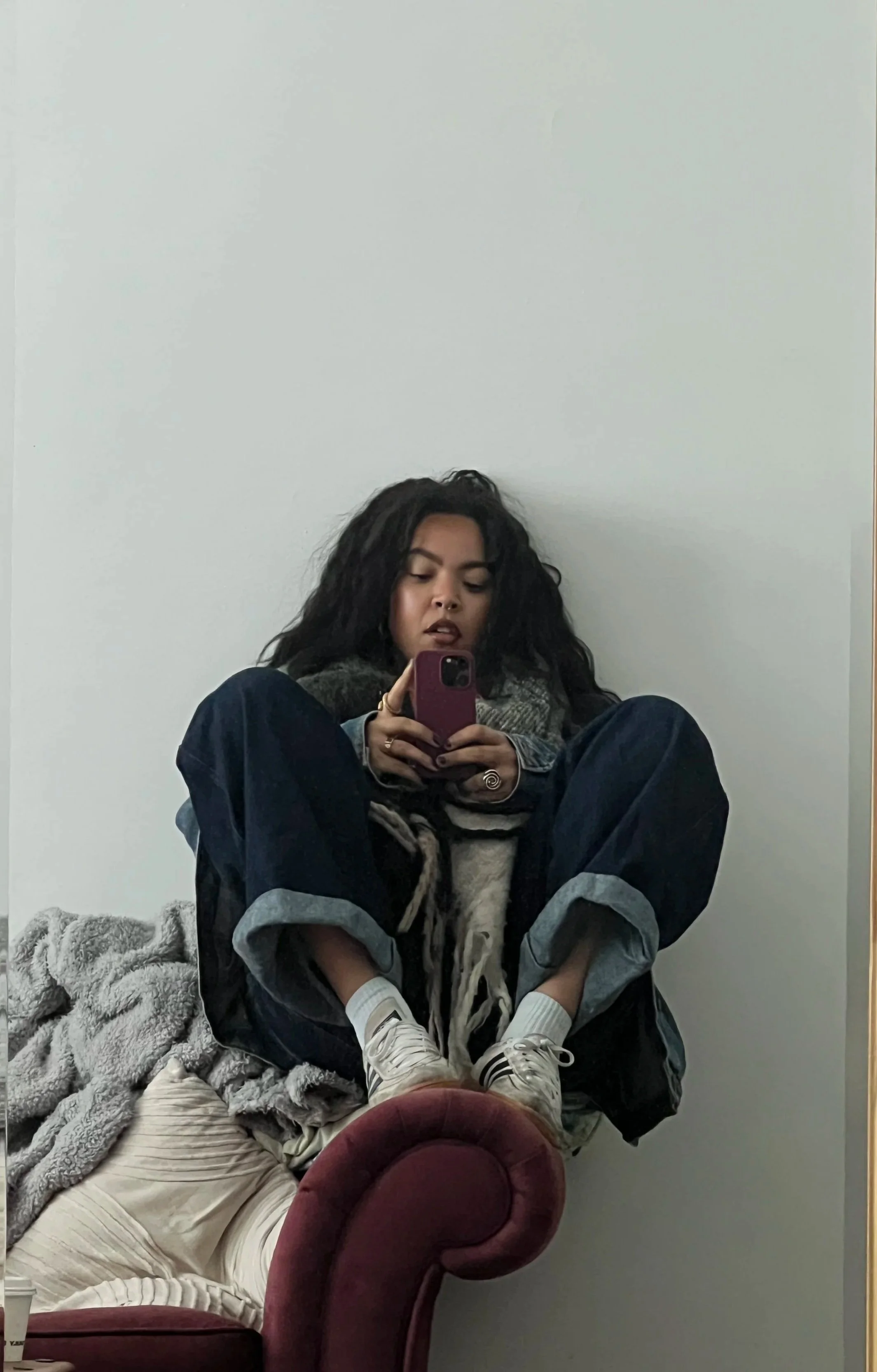 A woman sitting on a red velvet armchair, taking a selfie with her phone. She has curly dark hair, wearing a patterned jacket, white sneakers, and cream-colored pants. There is a blanket and some clothing on the chair next to her, against a plain white wall.