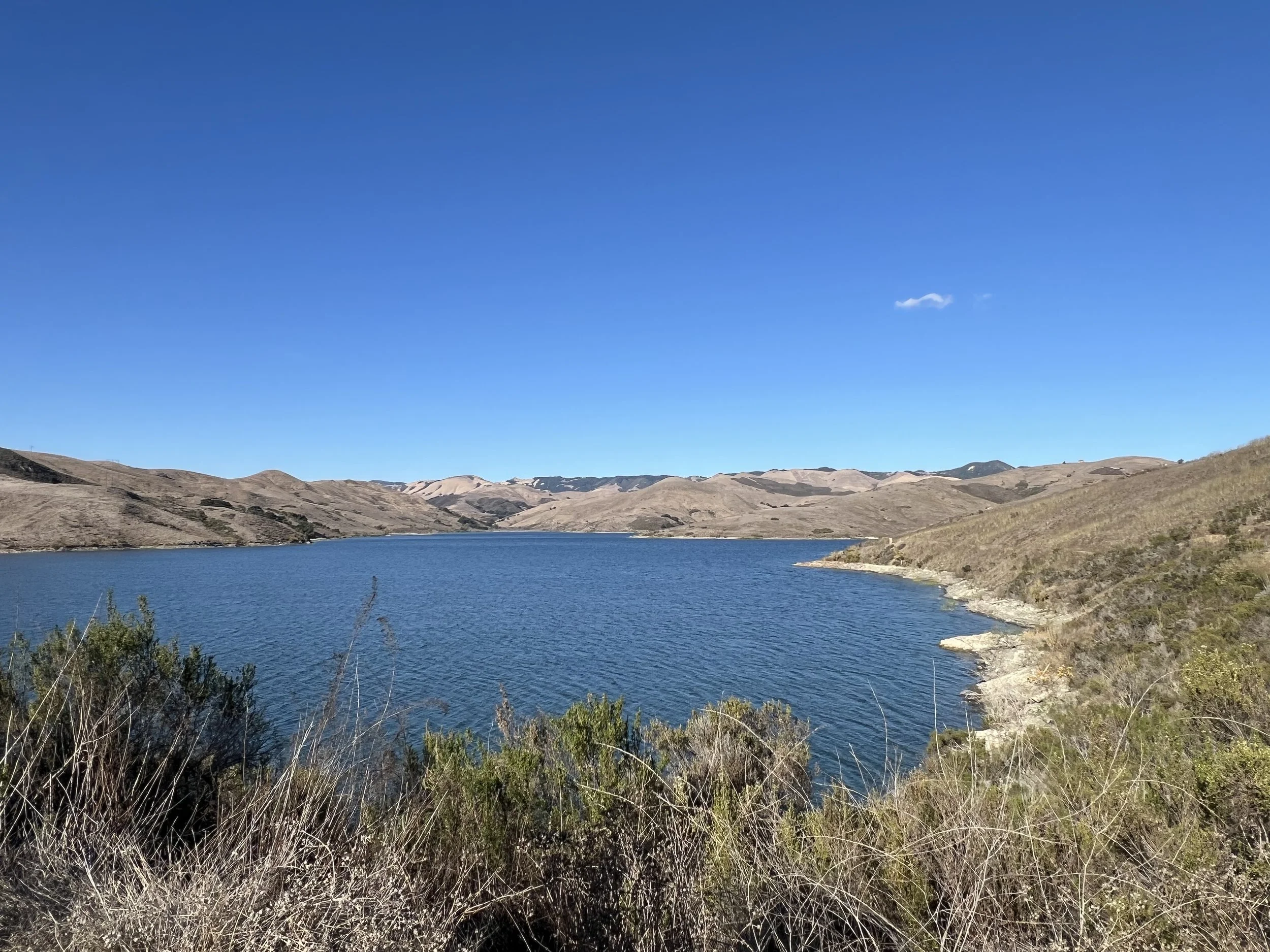 A landscape view of a lake with blue water surrounded by hills and dry grass under a clear blue sky.