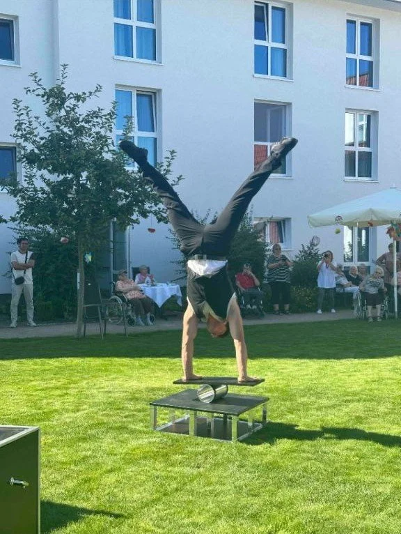 Mann macht Handstand auf einer runden Plattform im Garten, im Hintergrund Menschen bei einer Feier im Freien bei einem weißen Gebäude.