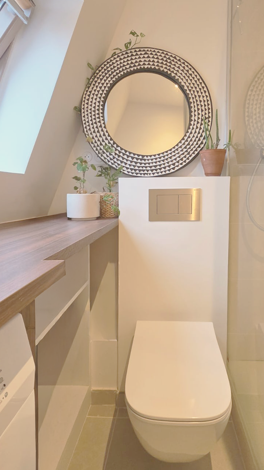 Toilettes blanches avec un panneau de commande encastré, miroir rond avec cadre noir et blanc, plantes en pot, et fenêtre à gauche dans une salle de bain moderne.