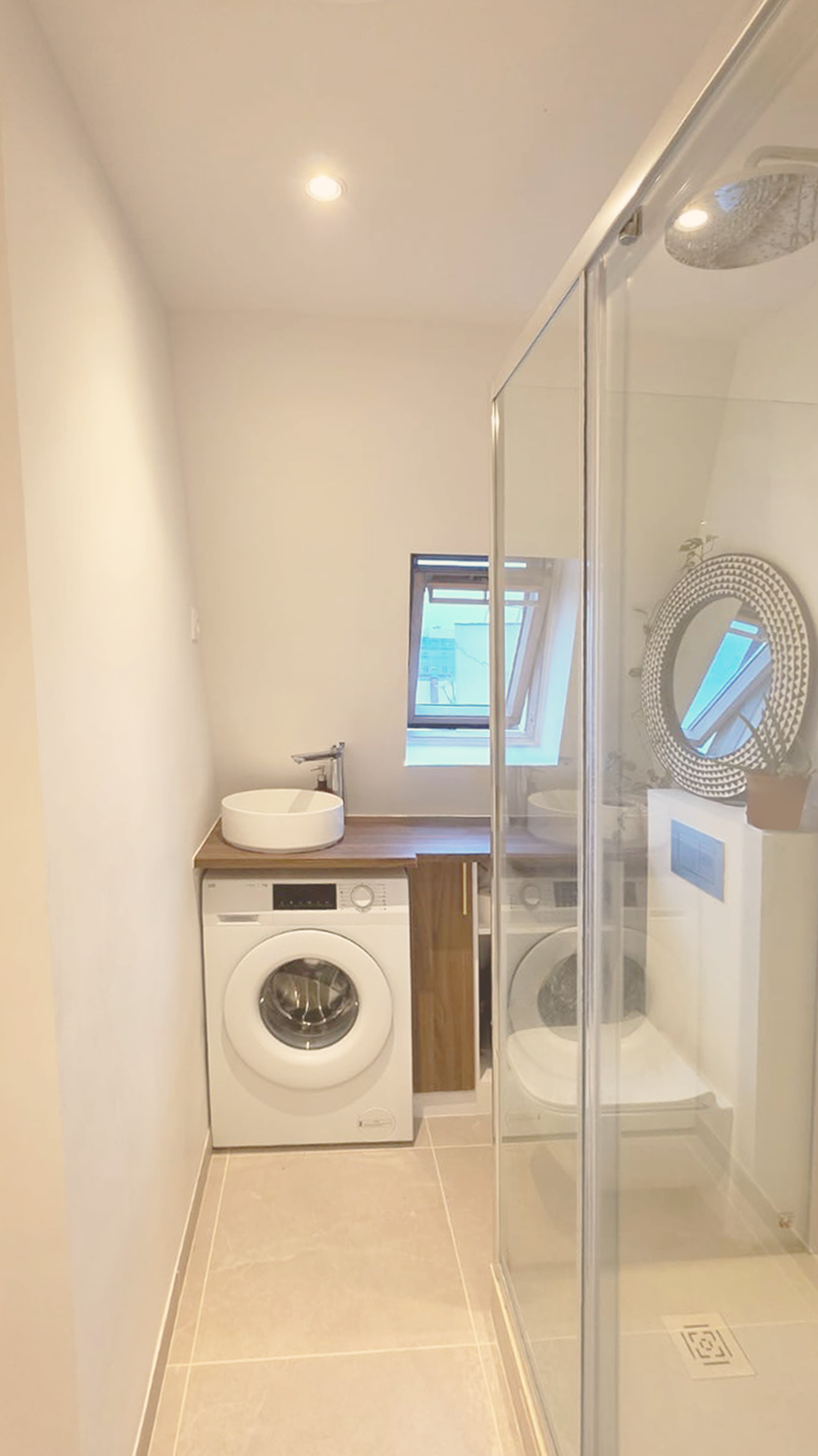 Une petite salle de bain avec une machine à laver, un lavabo rond sur un meuble en bois, une fenêtre inclinée laissant entrer de la lumière naturelle, et une cabine de douche en verre.