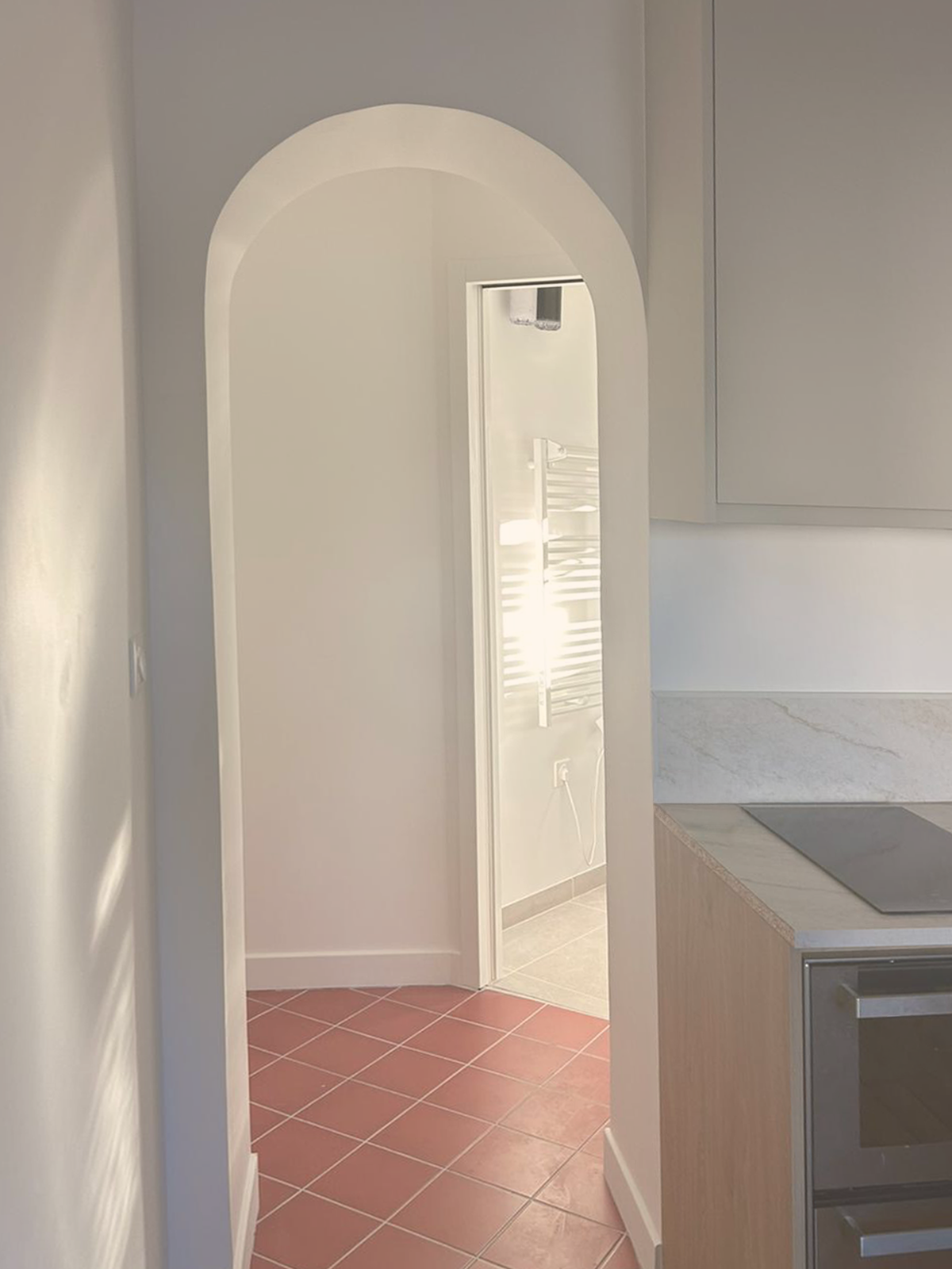 Intérieur d'une cuisine avec un comptoir en bois, une plaque de cuisson et un four, vue vers un couloir avec un sol en carreaux rouges.