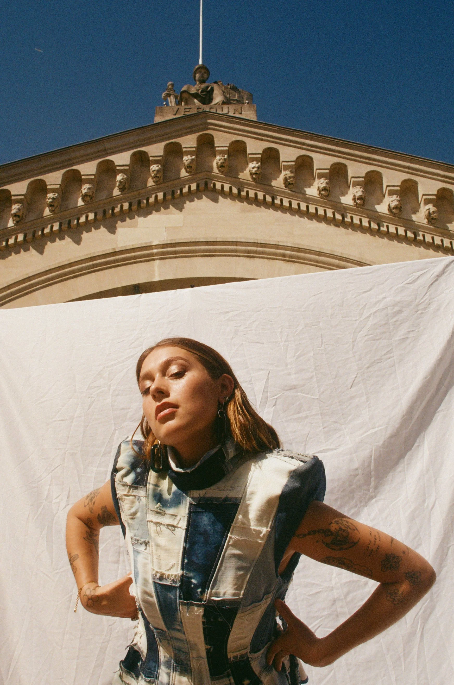 Une femme pose devant un fond blanc avec une architecture en pierre en arrière-plan, sous un ciel bleu clair.