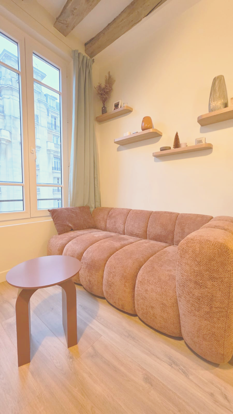 Un salon avec un canapé rose en tissu, une table basse marron, une fenêtre avec rideaux verticaux gris, et des étagères murales avec des décorations.