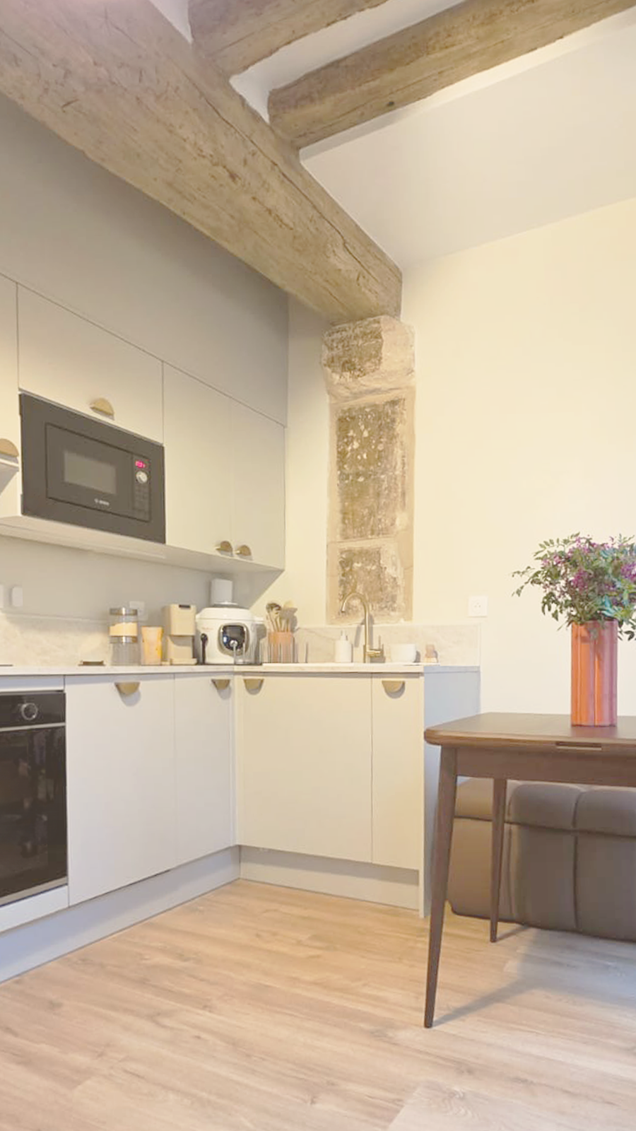 Cuisine moderne avec poutres en bois apparentes, armoires blanches, appareils électroménagers, table en bois et vase avec fleurs.