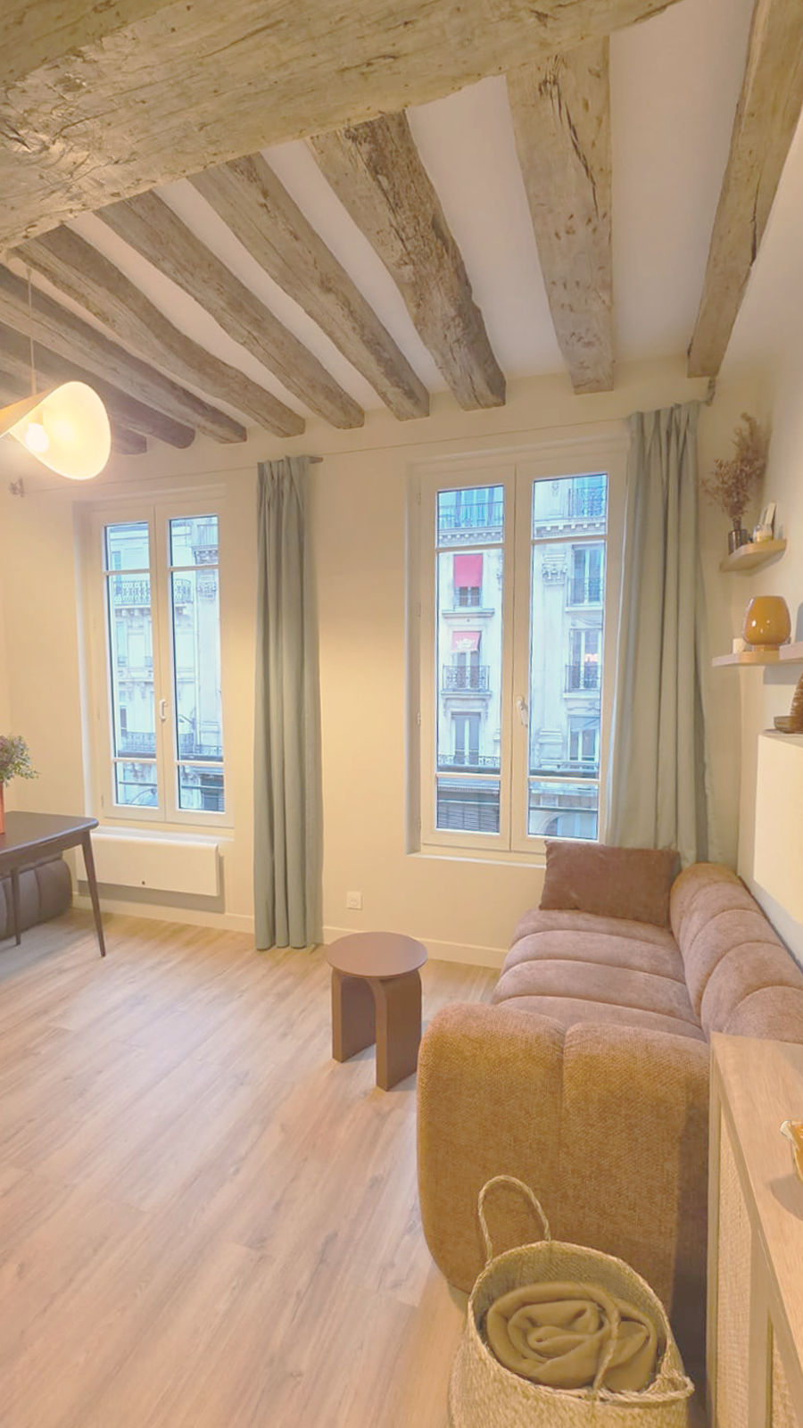 Salon avec canapé beige, petite table ronde, fenêtres avec rideaux verts, plafond en poutres en bois brut, décor minimaliste avec étagères flottantes et objets décoratifs.