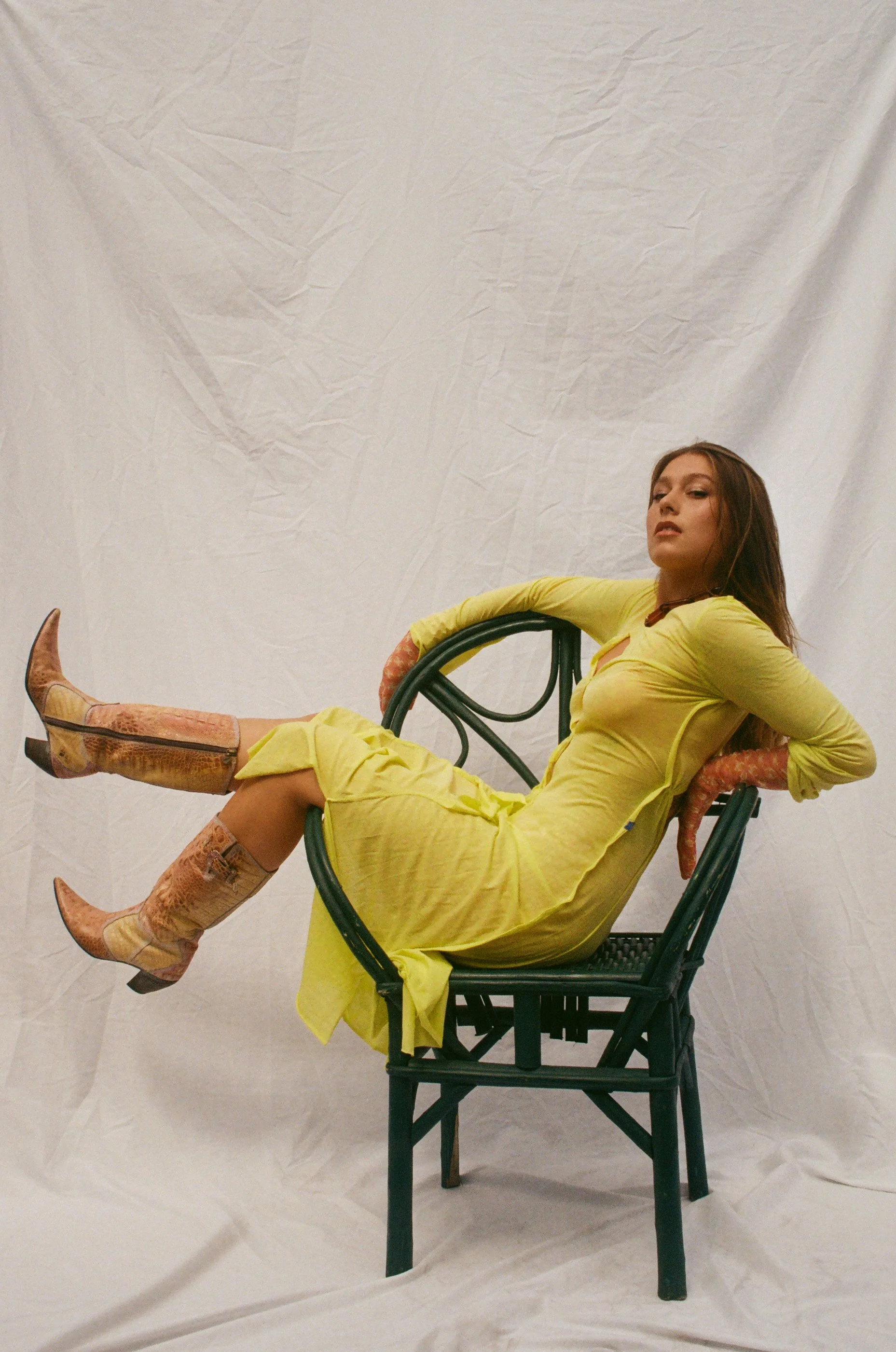 Femme en robe jaune à motifs, assise sur une chaise en rotin foncé, portant des bottes à motif serpent, devant un fond blanc