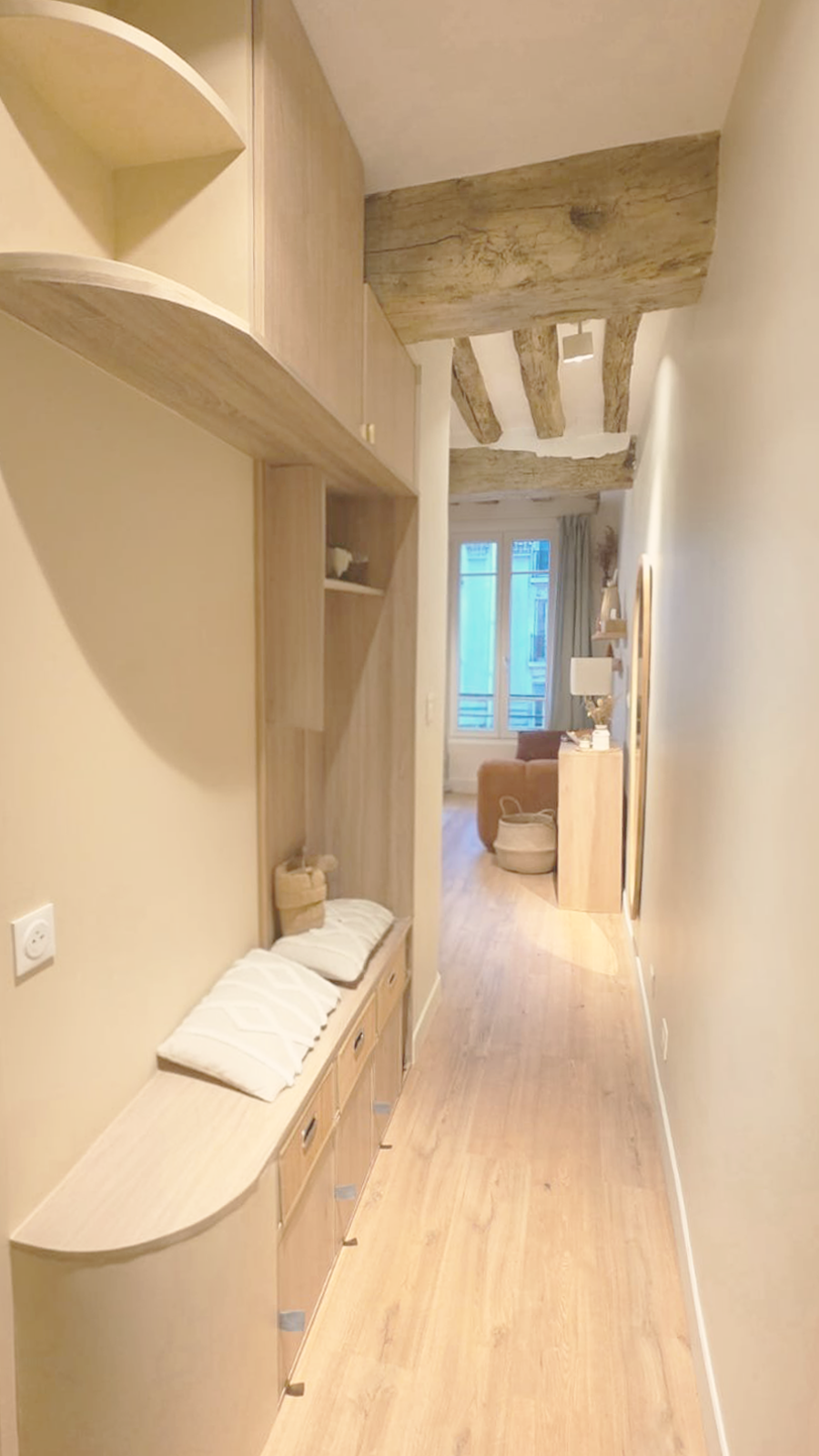 Un couloir dans un appartement avec des poutres en bois au plafond, un meuble en bois clair avec des coussins, et une pièce à vivre à l'arrière avec un fauteuil et une grande fenêtre.