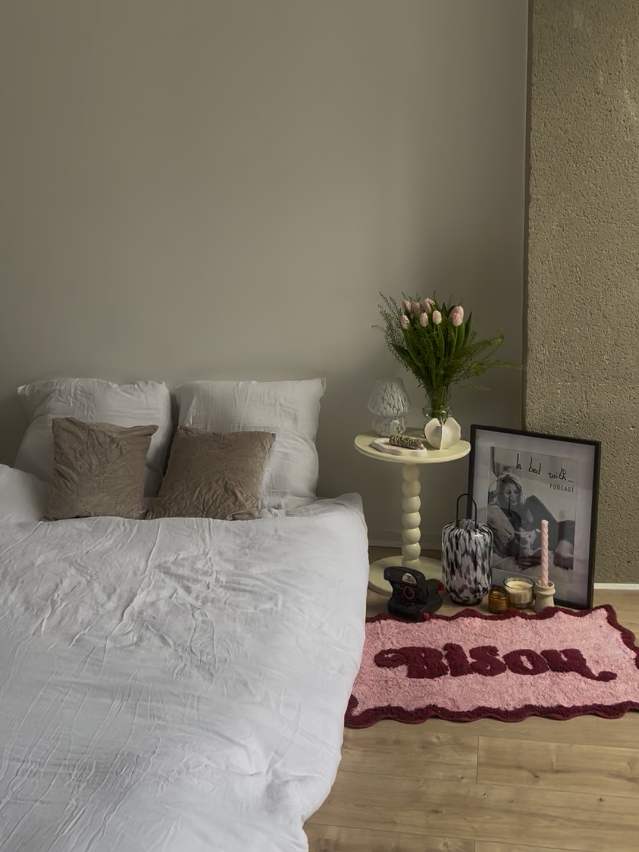 Chambre avec un lit blanc, deux coussins beige, table de chevet avec un vase de fleurs, cadre photo, bougies, et tapis rose