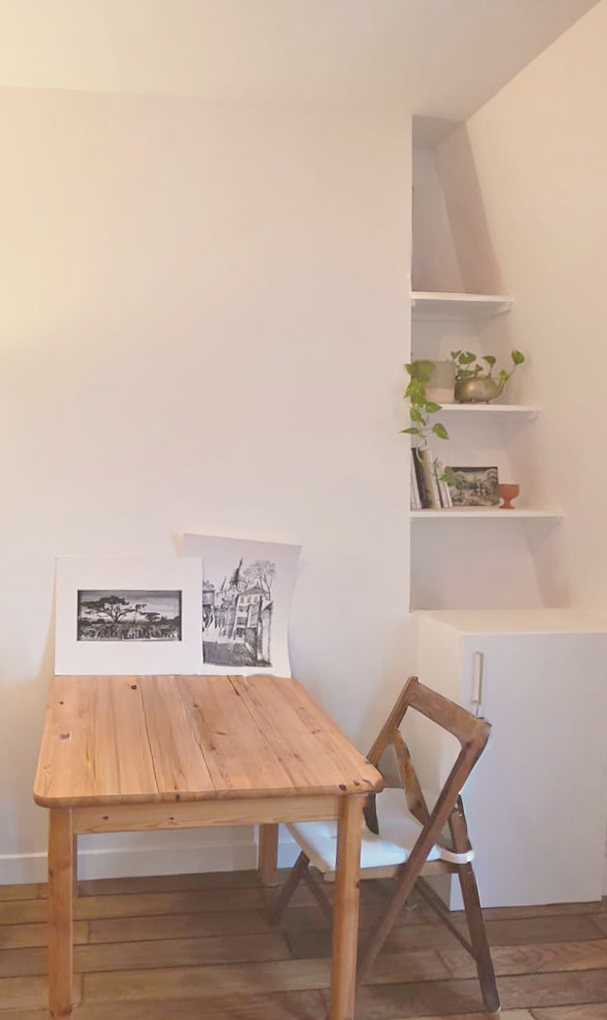 petit coin lecture avec un bureau en bois, une chaise pliante, et des étagères blanches avec des livres, des décorations en pot et une plante verte