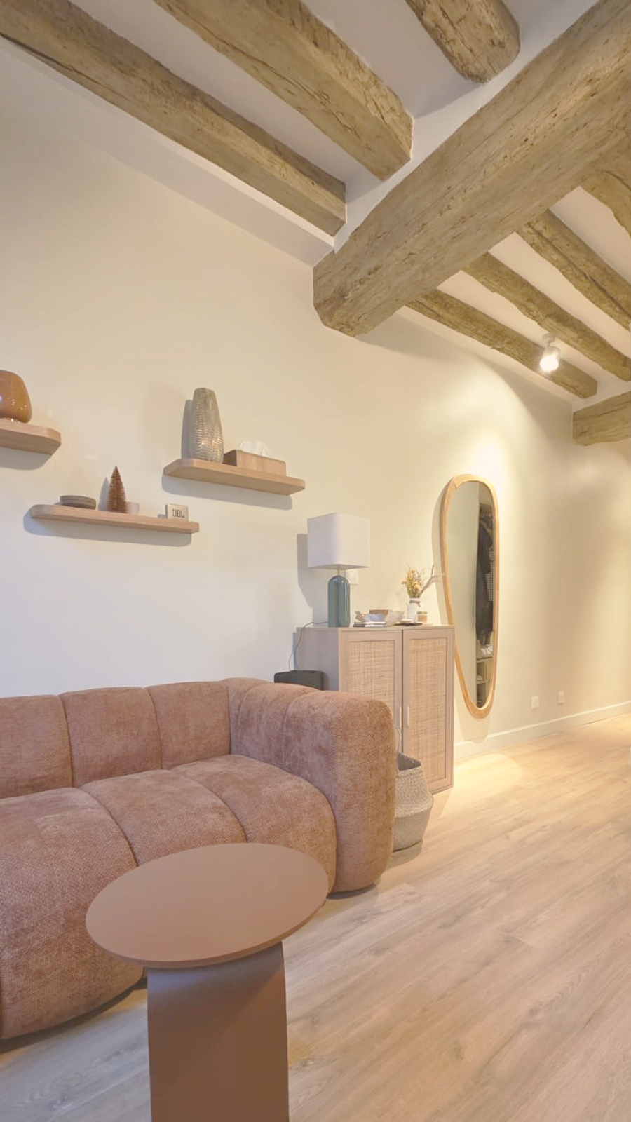Salon avec canapé beige, table d'appoint, meuble avec lampe, miroir ovale en bois, étagères avec décorations, plafond en poutres en bois.