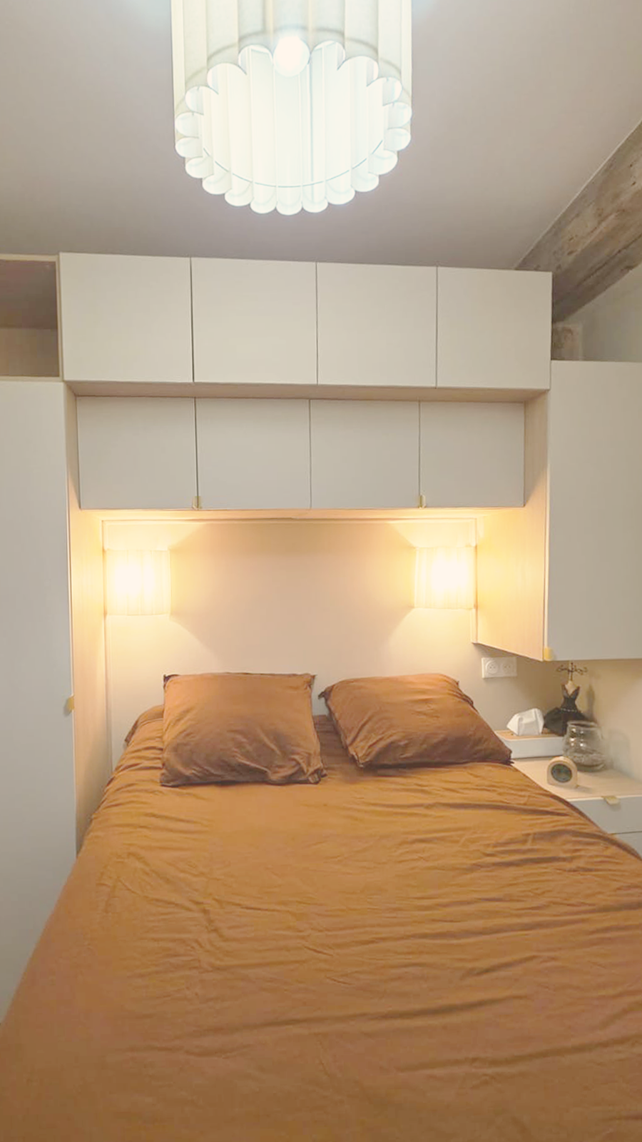 Lits avec oreillers en satin marron dans une chambre, éclairée par deux appliques murales, avec un plafond blanc et un luminaire blanc en haut.