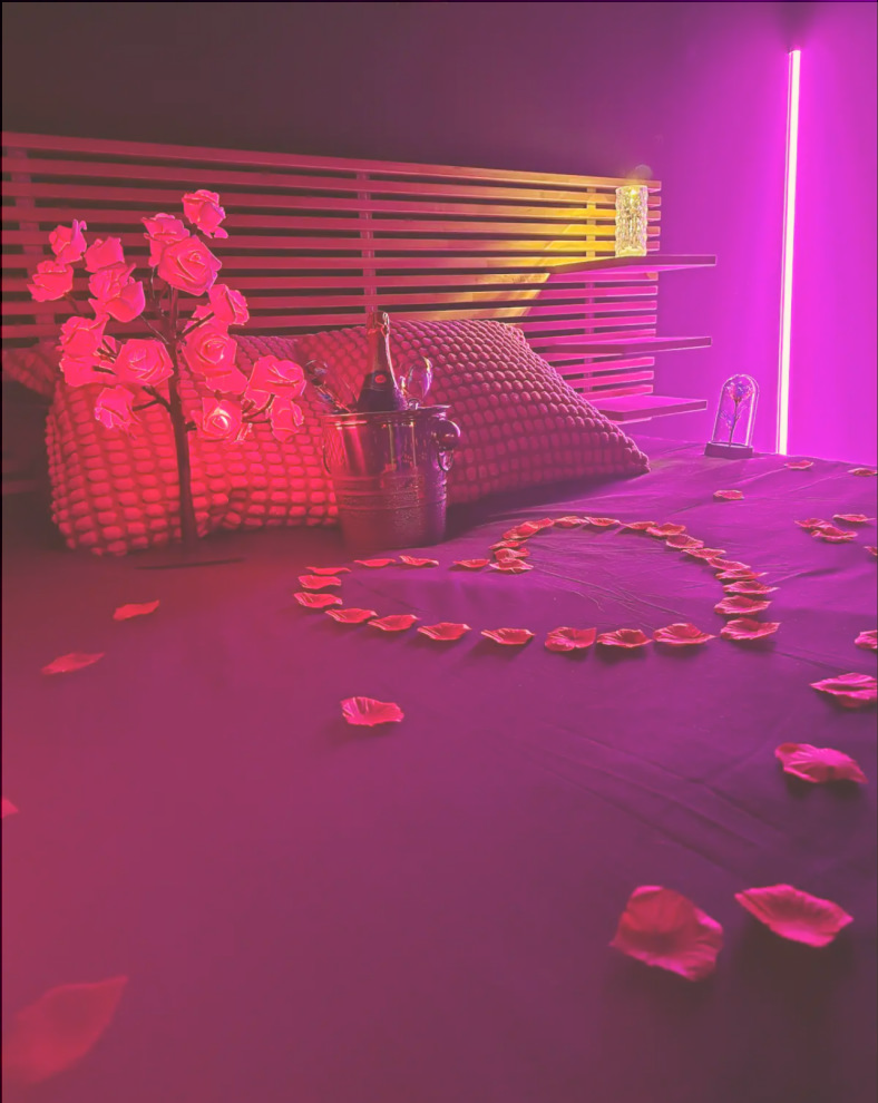 Lit romantique avec des roses en plastique, pétales de rose sur le lit, décoration roses et lumineuse en rose et violet.