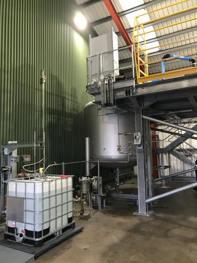 Industrial equipment with a large horizontal storage tank, connected to pipes and a smaller white water tank, set against a green corrugated metal wall in a factory or processing plant.
