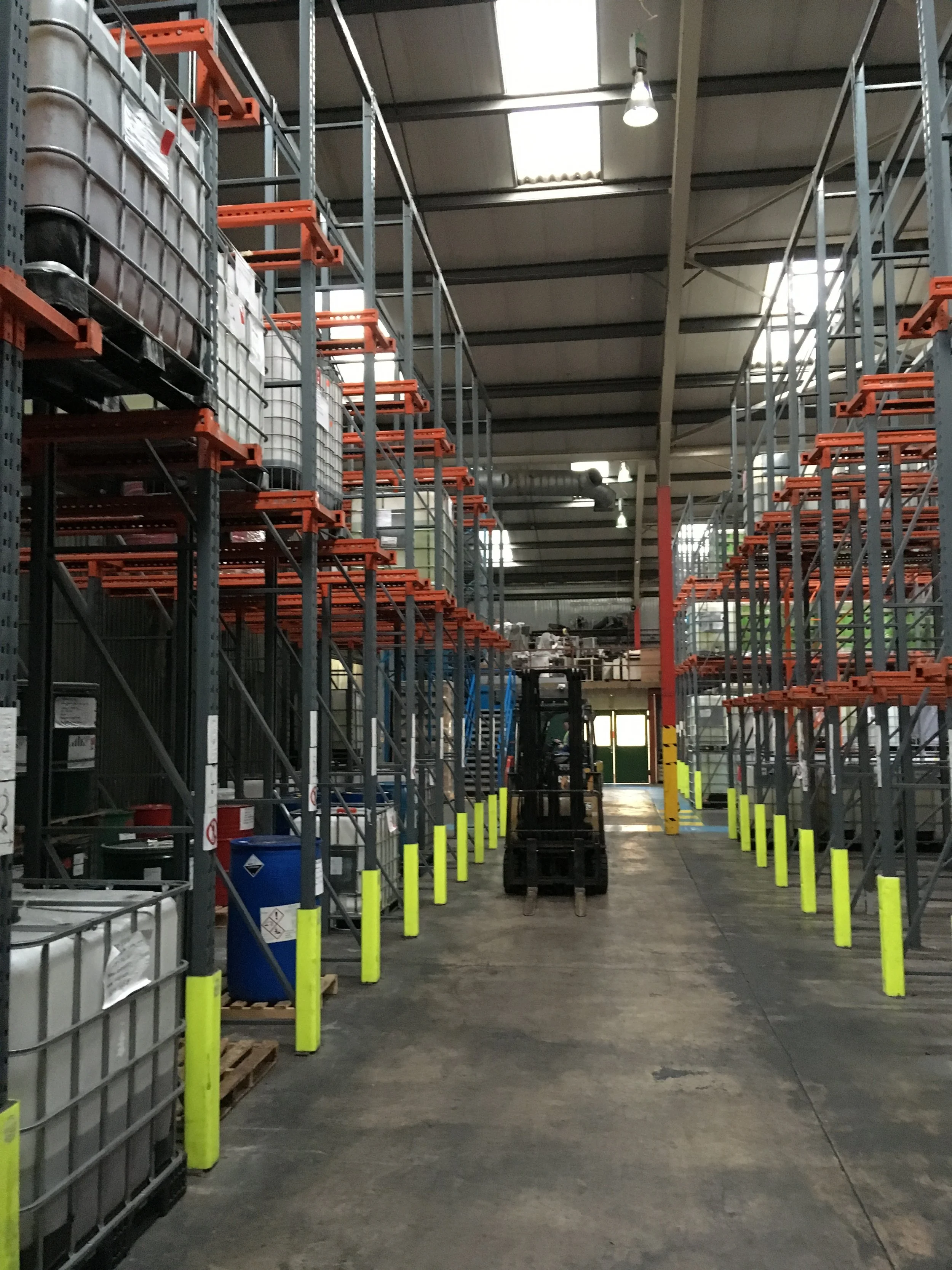 Industrial warehouse aisle with metal shelving racks on both sides, some with large containers. A forklift is parked in the center and yellow protective barriers line the aisle.