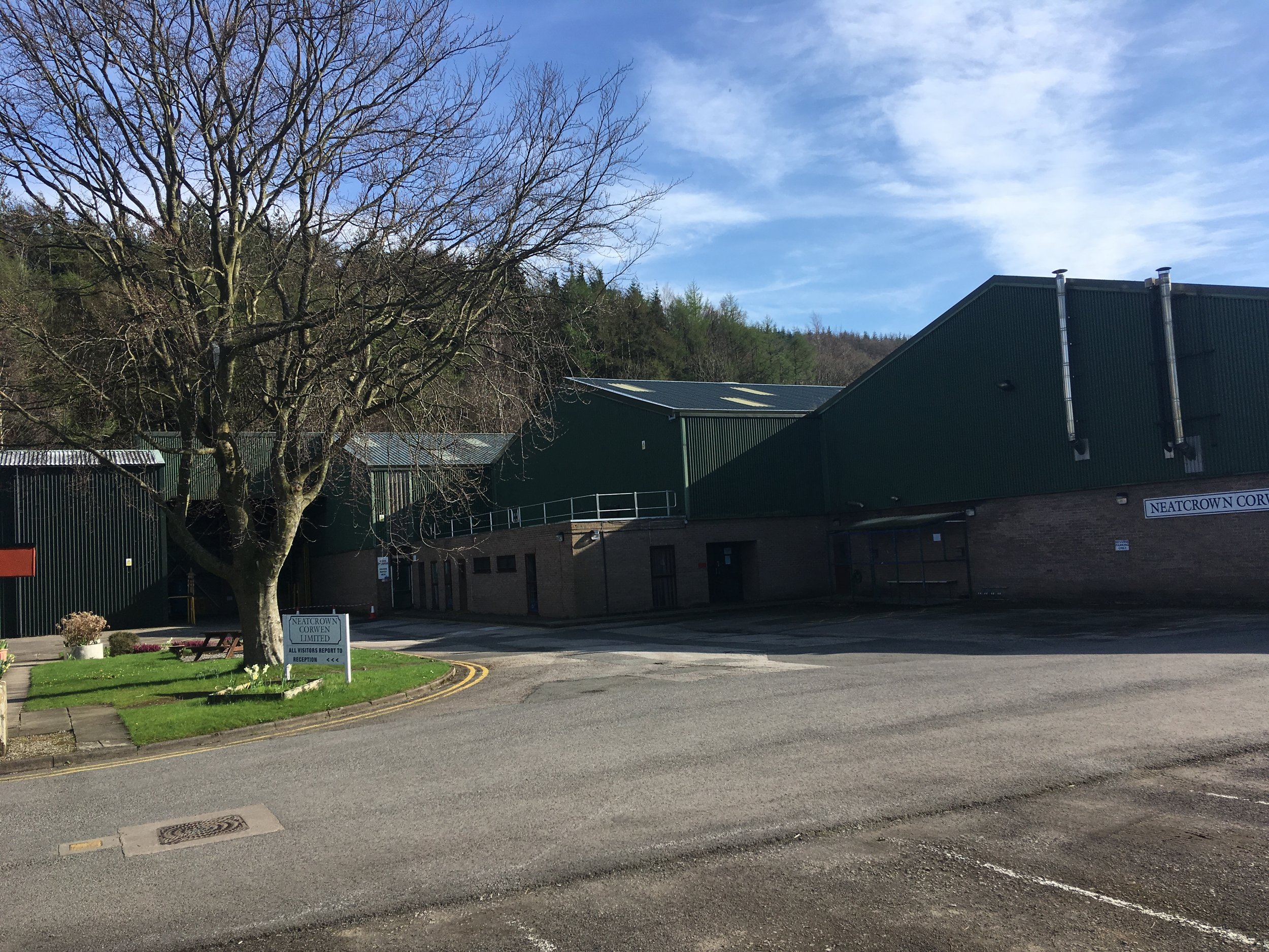 Exterior view of a building with green siding, brick lower walls, and a large leafless tree in front. A sign indicates 'Neatcrown Corwen Limited' at the entrance, with a parking area and a hill covered with trees in the background under a partly clou