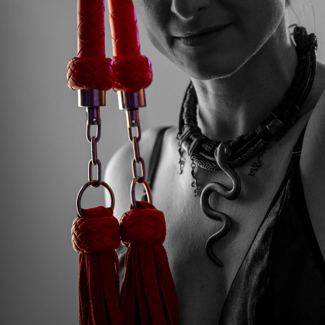 A person holding a pair of red martial arts or boxing hand wraps with metallic clips, wearing a black leather top and a black snake-shaped necklace. The image is primarily in black and white with the hand wraps in color.