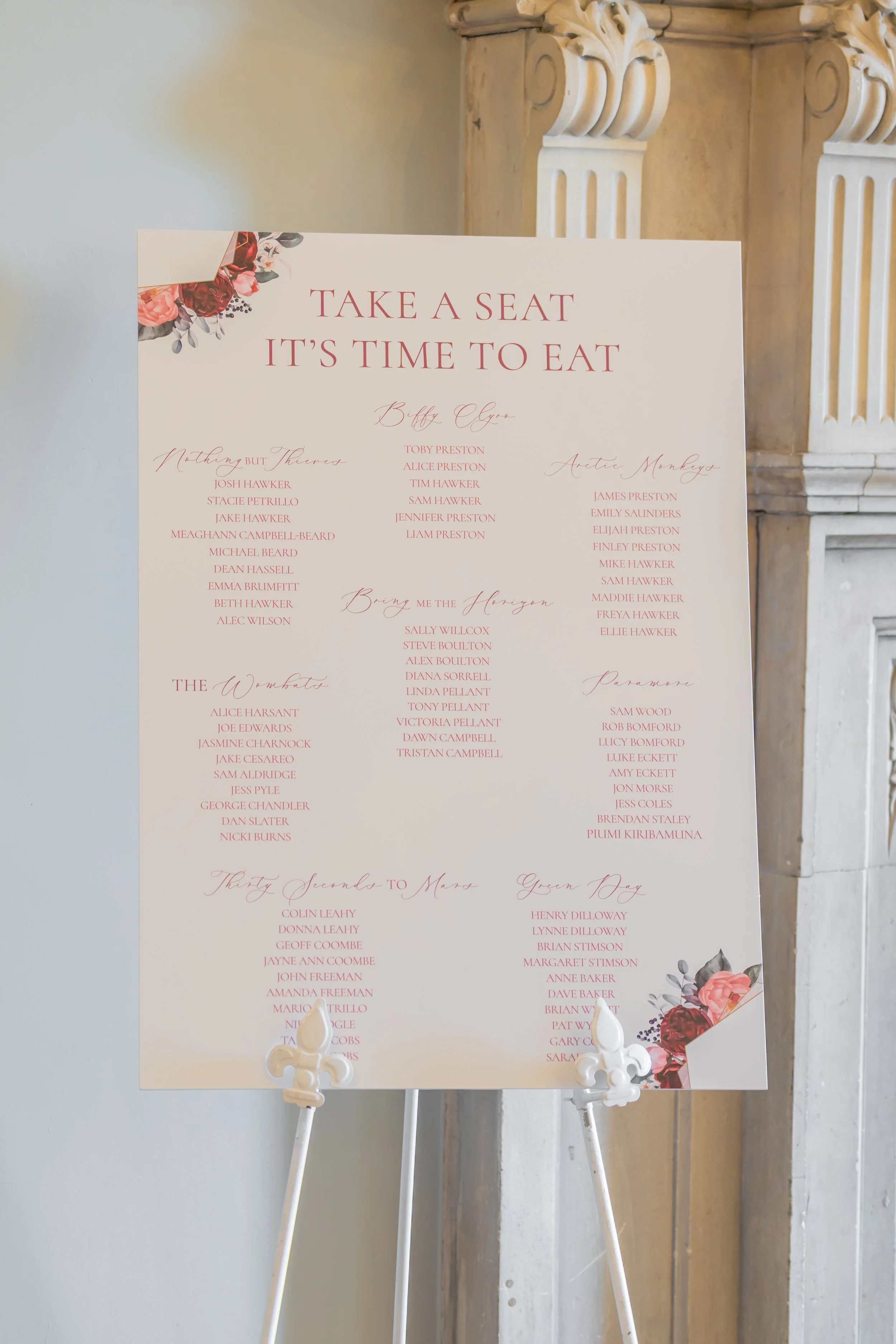 A wedding seating chart on a white easel with floral decorations in the corners, listing guests' names under various sections.