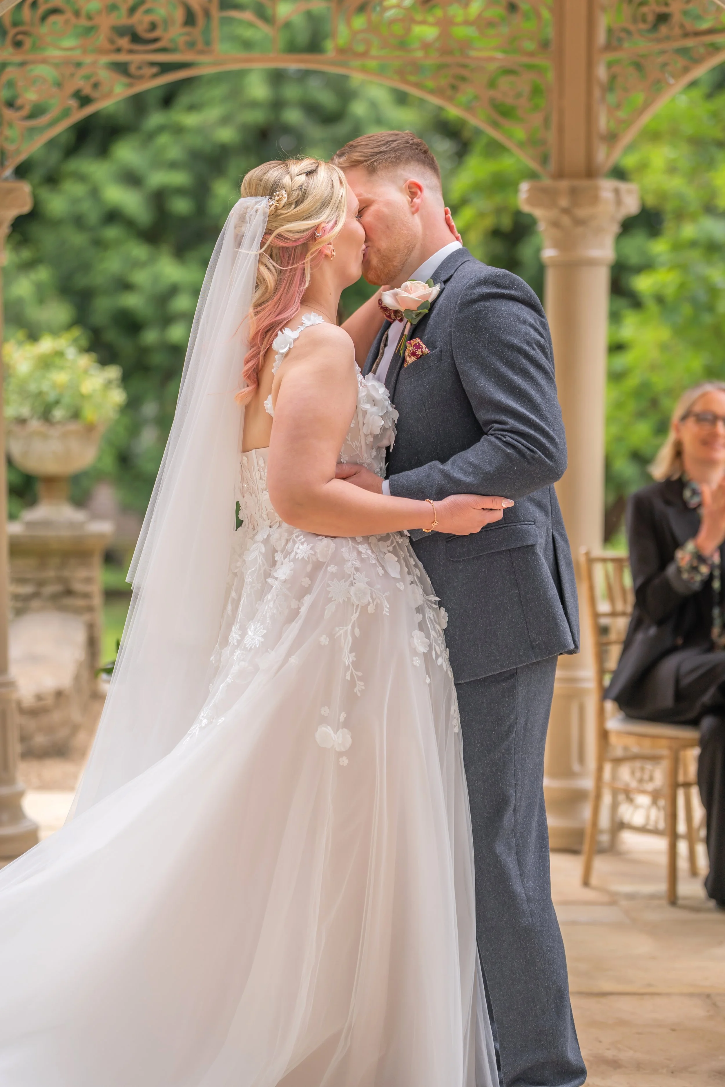 A bride and groom kiss during their outdoor wedding ceremony, with wedding officiant in the background.