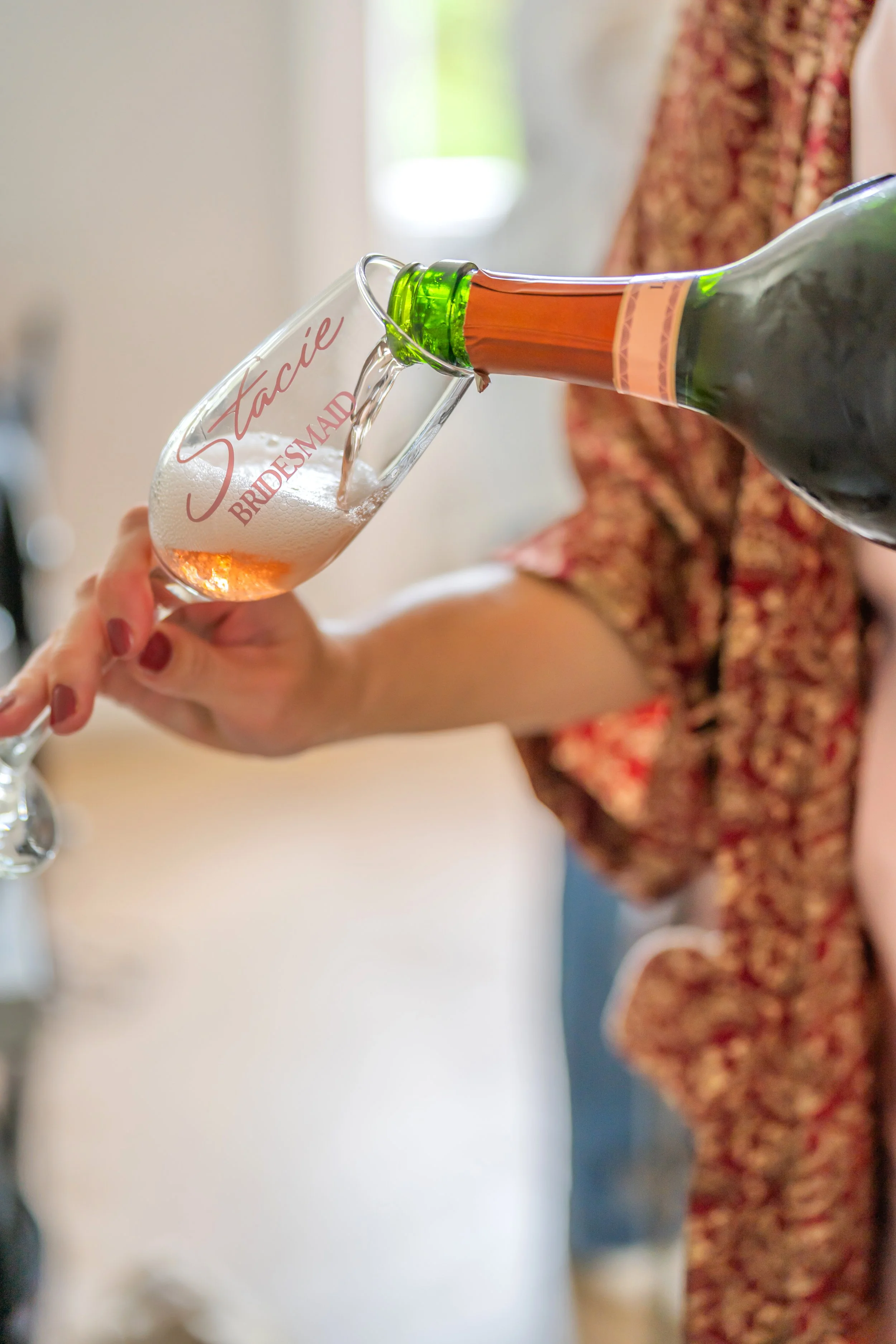 A woman in a floral dress is pouring champagne from a bottle into a branded flute glass that says 'Stacie Bridesmaid'.