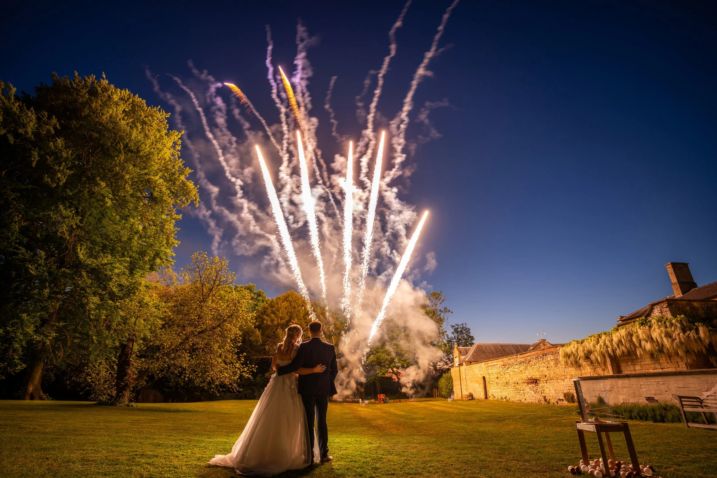 A bride and groom watching fireworks over a field at night, with trees on the left and a stone building on the right.