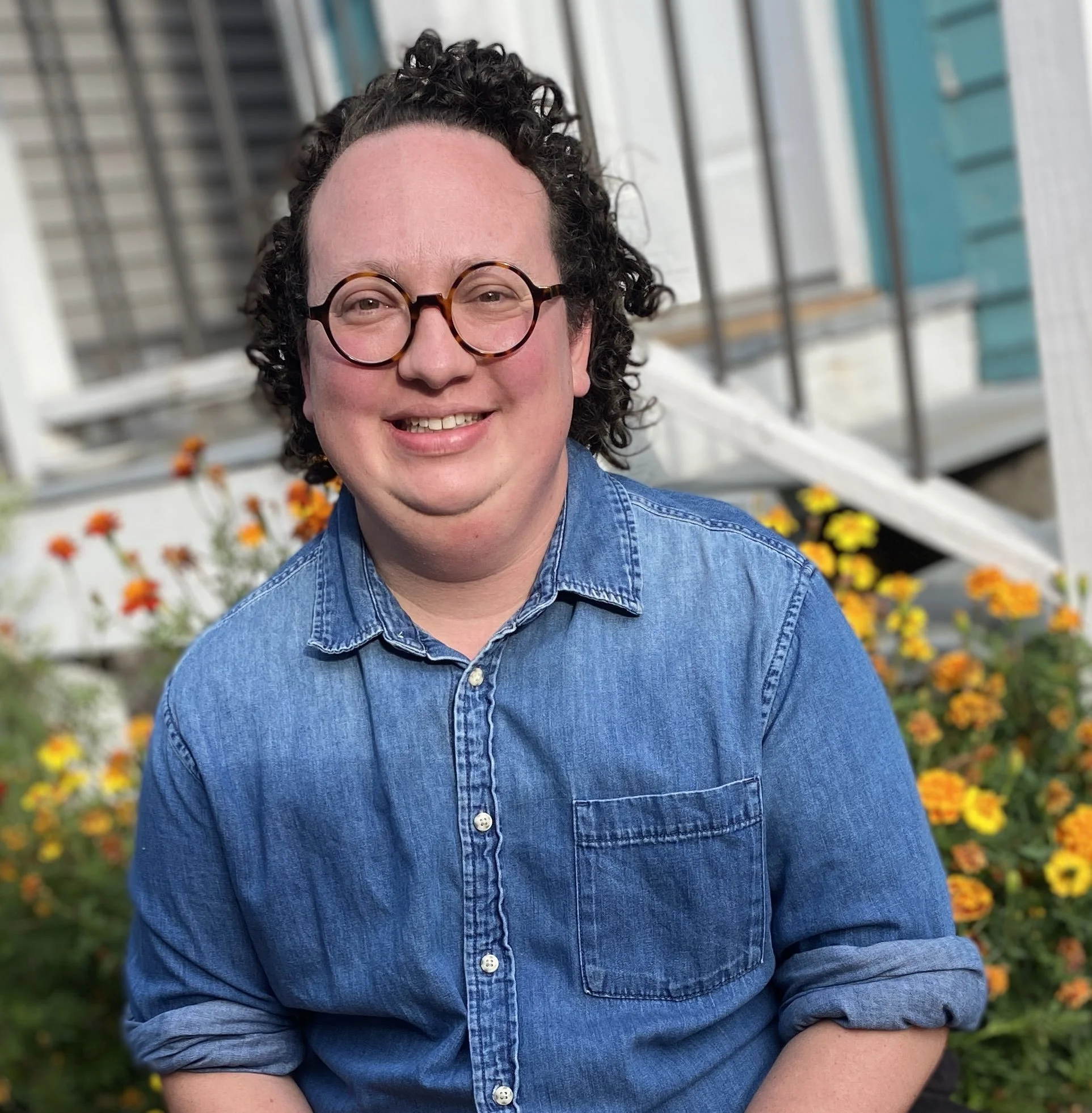 August Kalinosky, Minnesota Death Doula, sitting with orange mariolds and a teal and white house behind them. They have light skin, dark curly hair, round glasses, and a denim shirt.