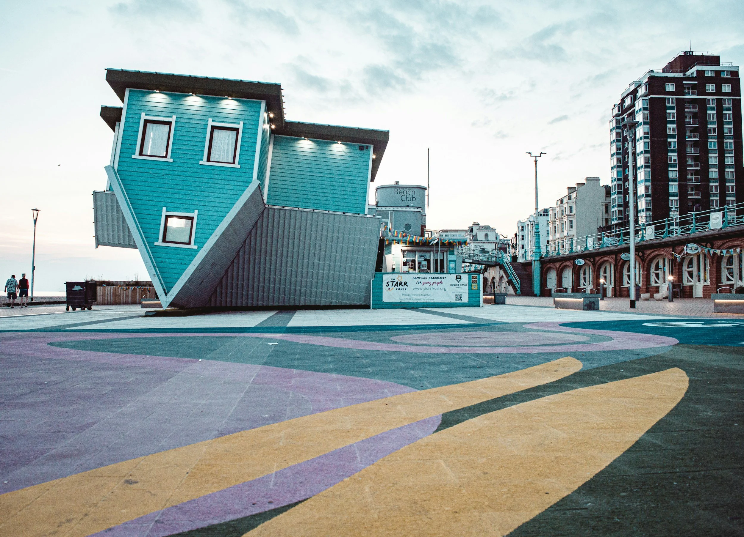 A colorful, geometric sidewalk pattern with curved lines in various pastel shades. In the background, a curved building with a water tower labeled 'Beach Club' and several high-rise buildings.
