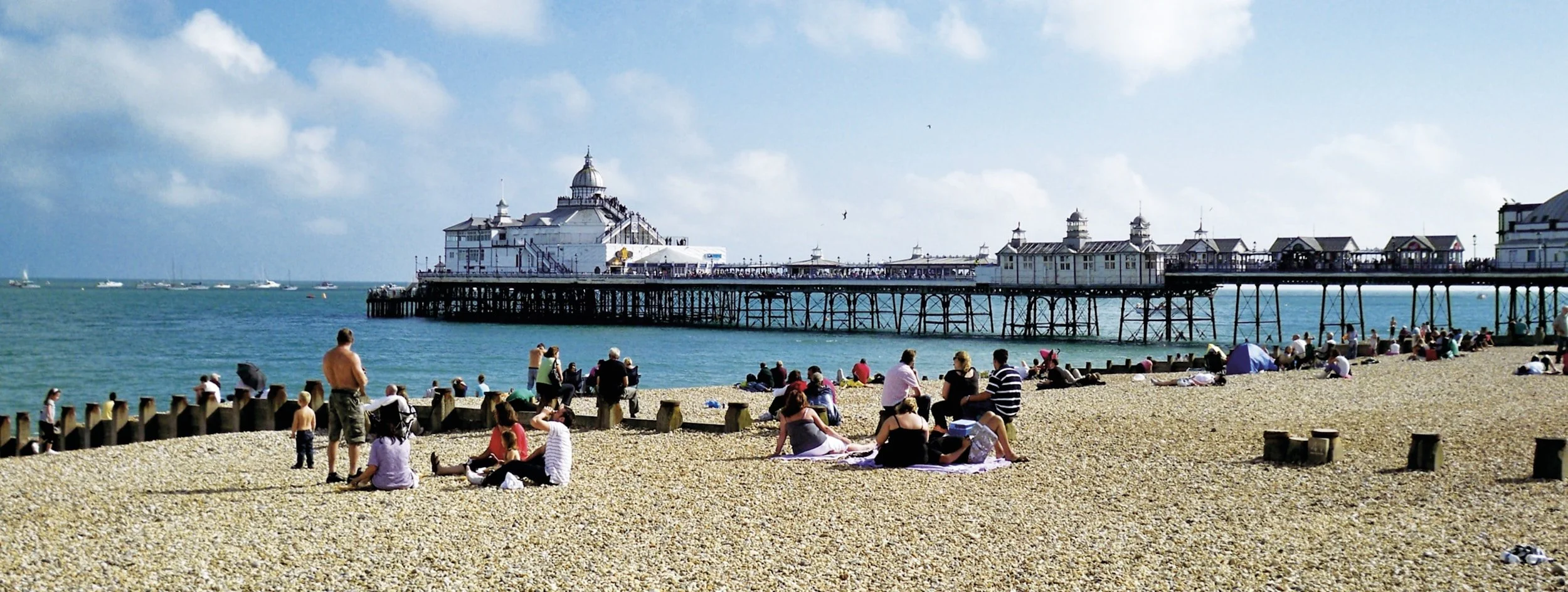 People relaxing on a pebble beach near a pier with a large building at the end, under a partly cloudy sky.
