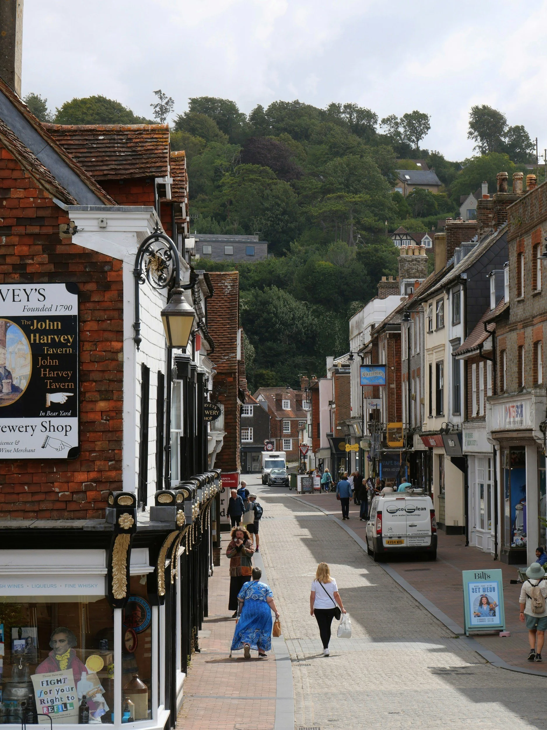 A street scene in a small town featuring shops and pedestrians, with a hillside covered in trees in the background.