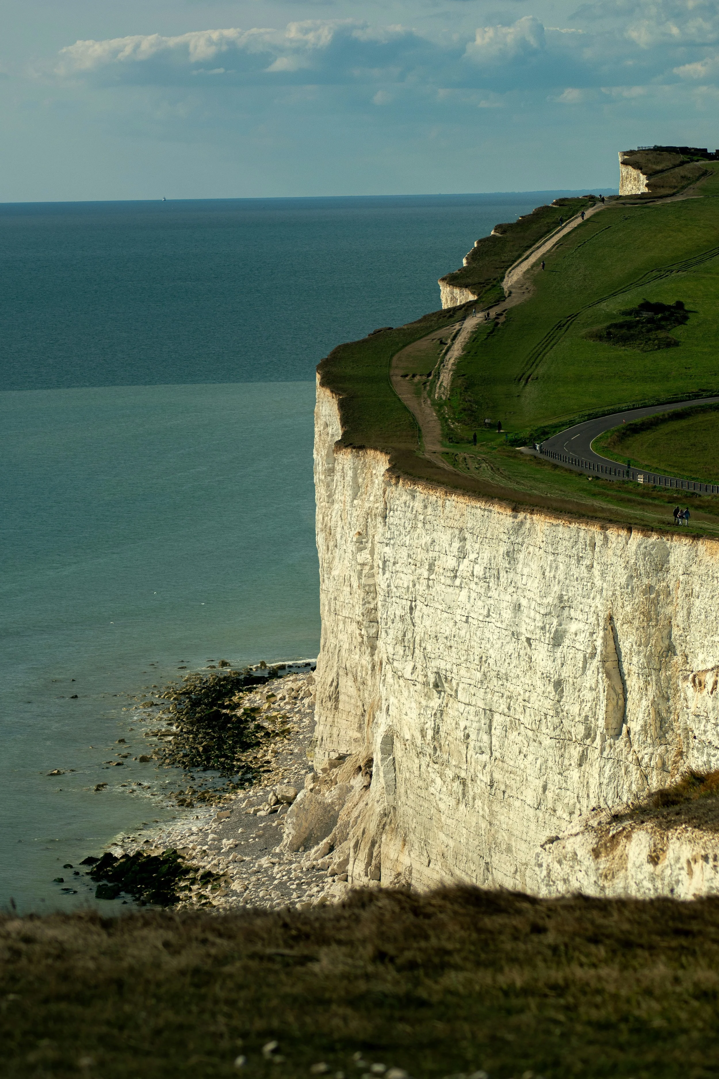 Cliffs along the coast with grassy areas and walking paths, overlooking the ocean with a sailboat in the distance.