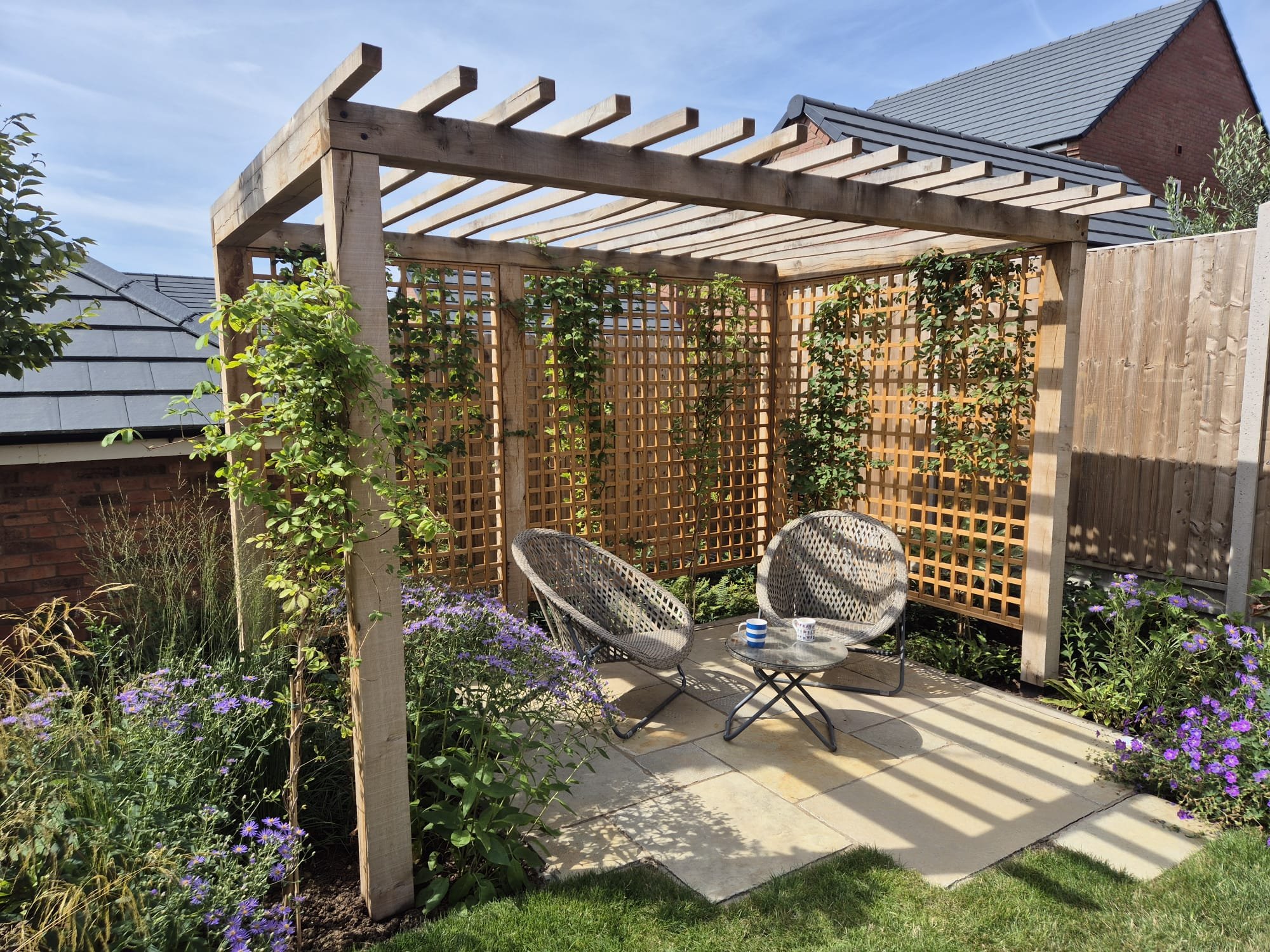 A backyard patio area with a wooden pergola and lattice panels, surrounded by purple flowers and green foliage. There are two woven chairs and a small round table with mugs on it, on a concrete patio.