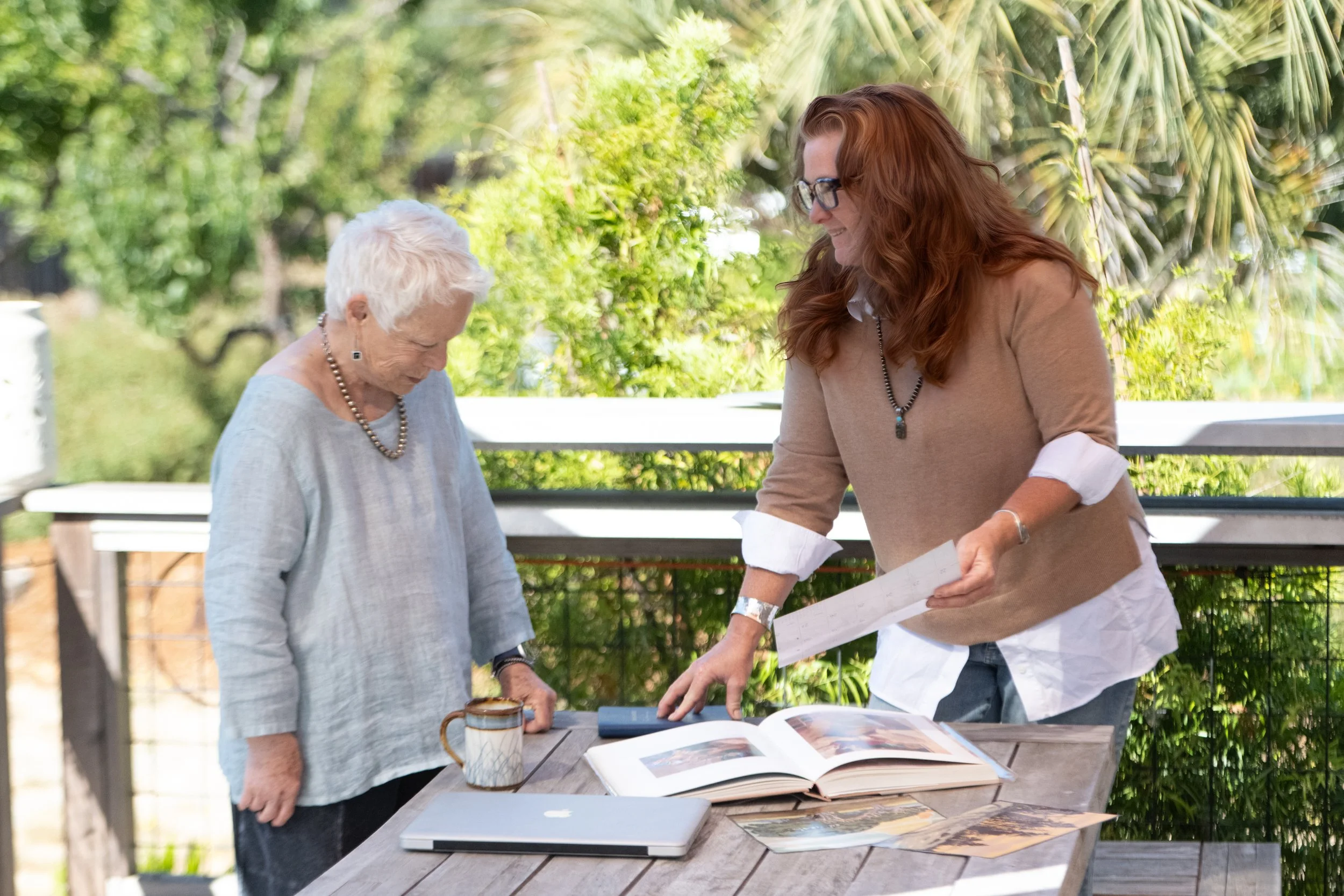 Two women standing by a table outdoors, looking at photo albums and papers, with greenery in the background.