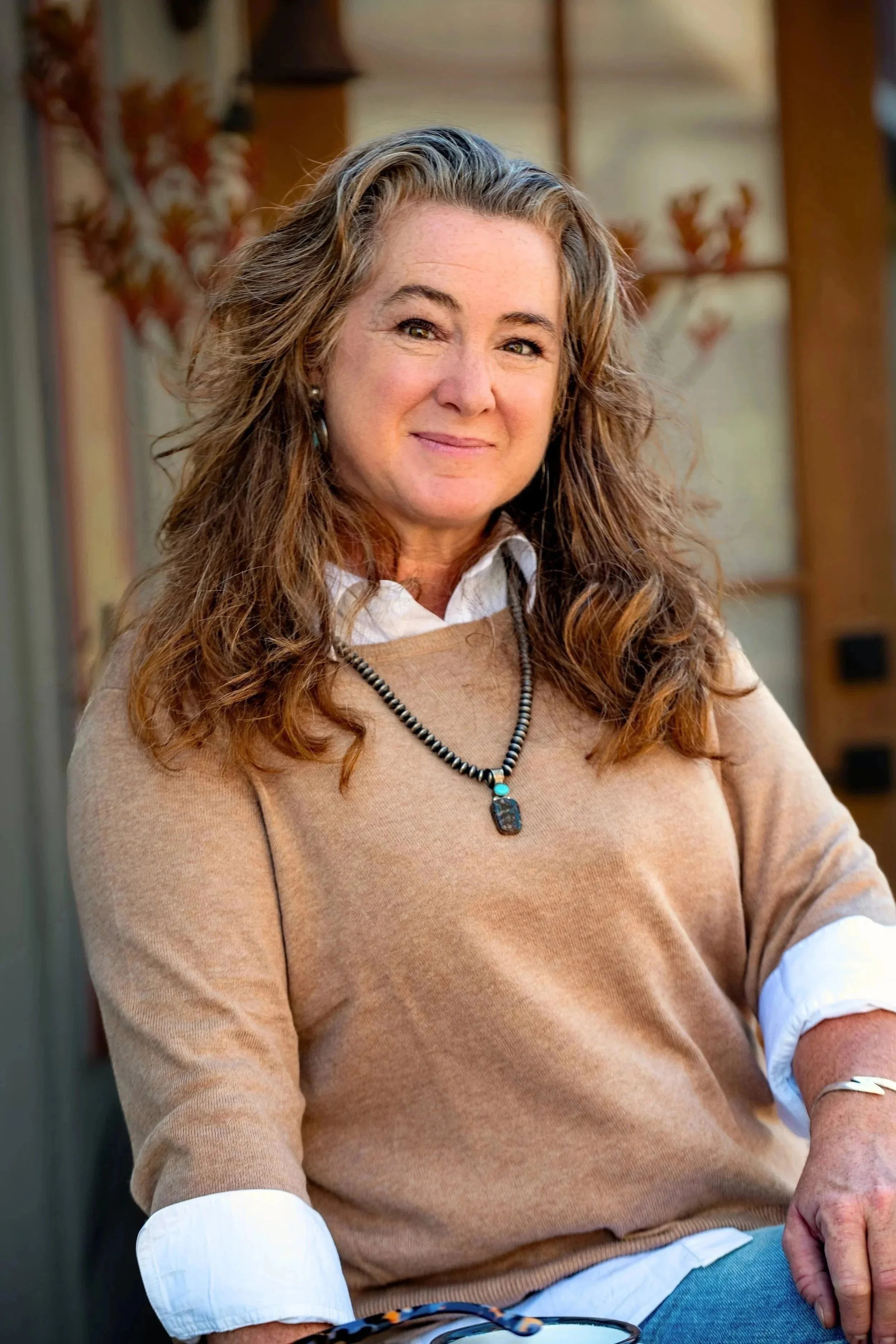 A woman with long, wavy, light brown hair, wearing a white shirt under a tan sweater, sitting outdoors with a blurred background of autumn leaves and wooden structures, smiling slightly at the camera.