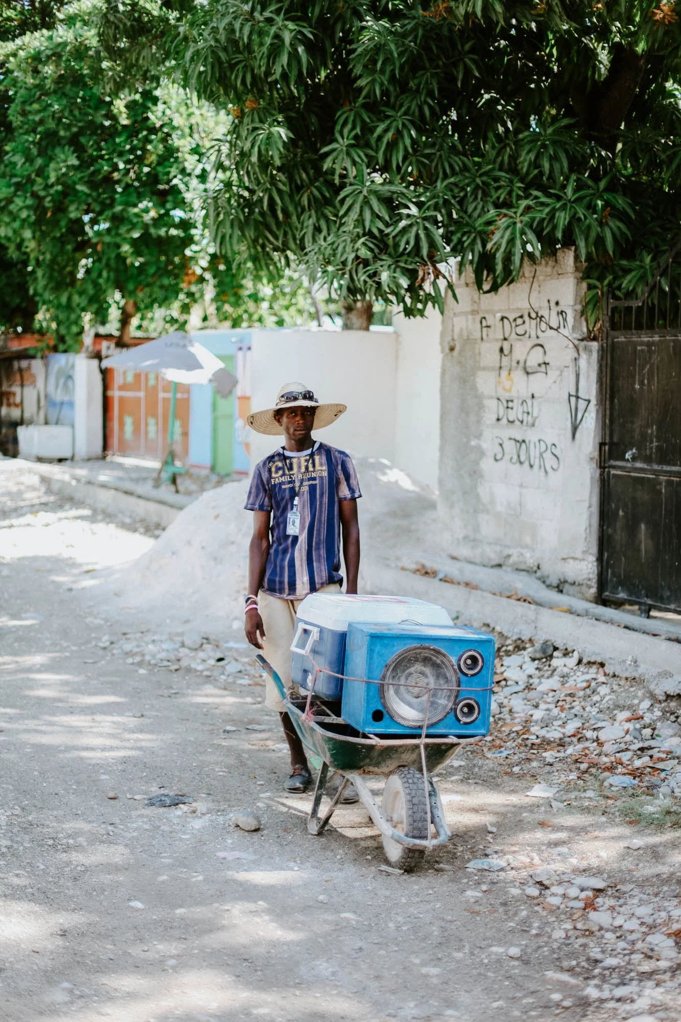A man wearing a large straw hat and sunglasses pushes a wheelbarrow containing a speaker and cooler along a dirt road shaded by green trees.